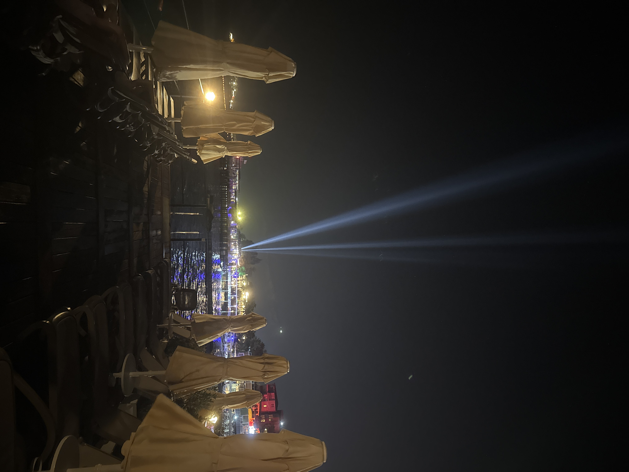 The image depicts a nighttime scene on a pier in Kemer, Turkey. Several closed beach umbrellas are arranged in rows on a wooden deck, with lounge chairs partially visible beneath them. In the background, a body of water is illuminated by vibrant lights from buildings and floating structures. Two powerful beams of light cut through the dark night sky from the distance, projecting upwards. The overall ambiance suggests a resort or entertainment area.