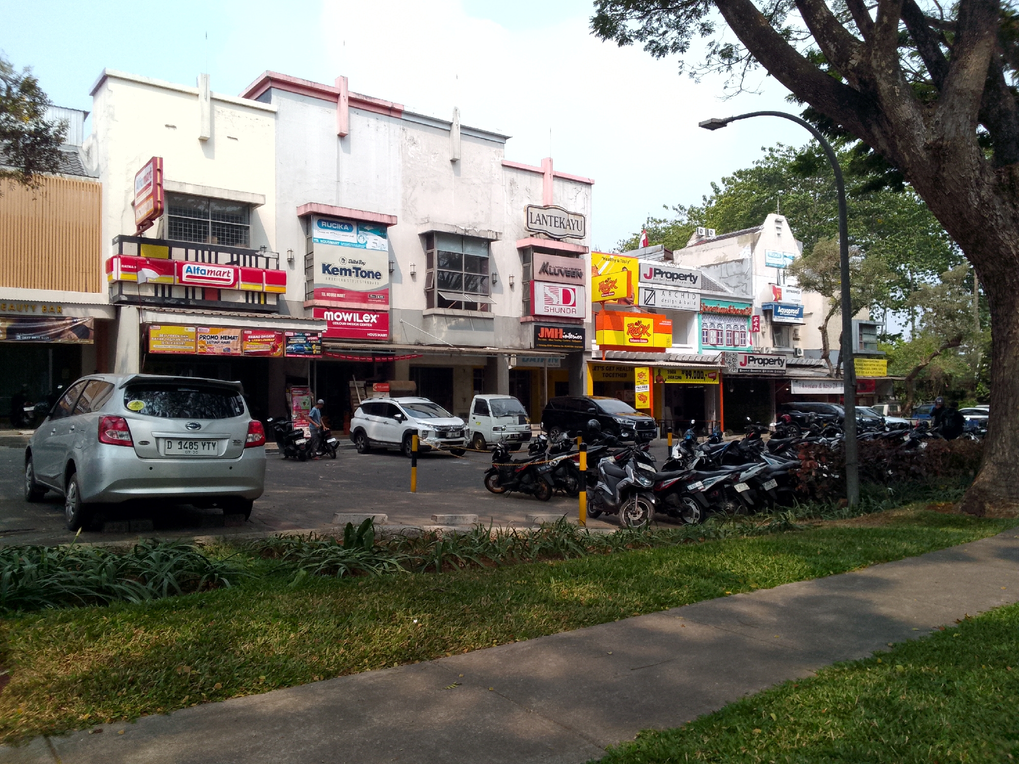 The image shows a street scene in Padalarang, Indonesia, featuring a row of commercial buildings with various storefronts. On the left, an Alfamart convenience store is visible, with its red and yellow signage prominently displayed. Adjacent to it, other businesses include RUCIA, Kem-Tone, and Mowilex. Further down the street, signs for LANTEKAYU, J PROPERTY, and ROCK are present. A silver car with license plate D 1485 VTY is parked in front of the buildings. Several motorcycles are parked in a designated area, with some people interacting near them. The buildings have a mix of architectural styles, with some displaying older facades and others with more modern signage. Trees and greenery line the sidewalk, and a street lamp is visible. The sky is clear, suggesting a daytime scene.