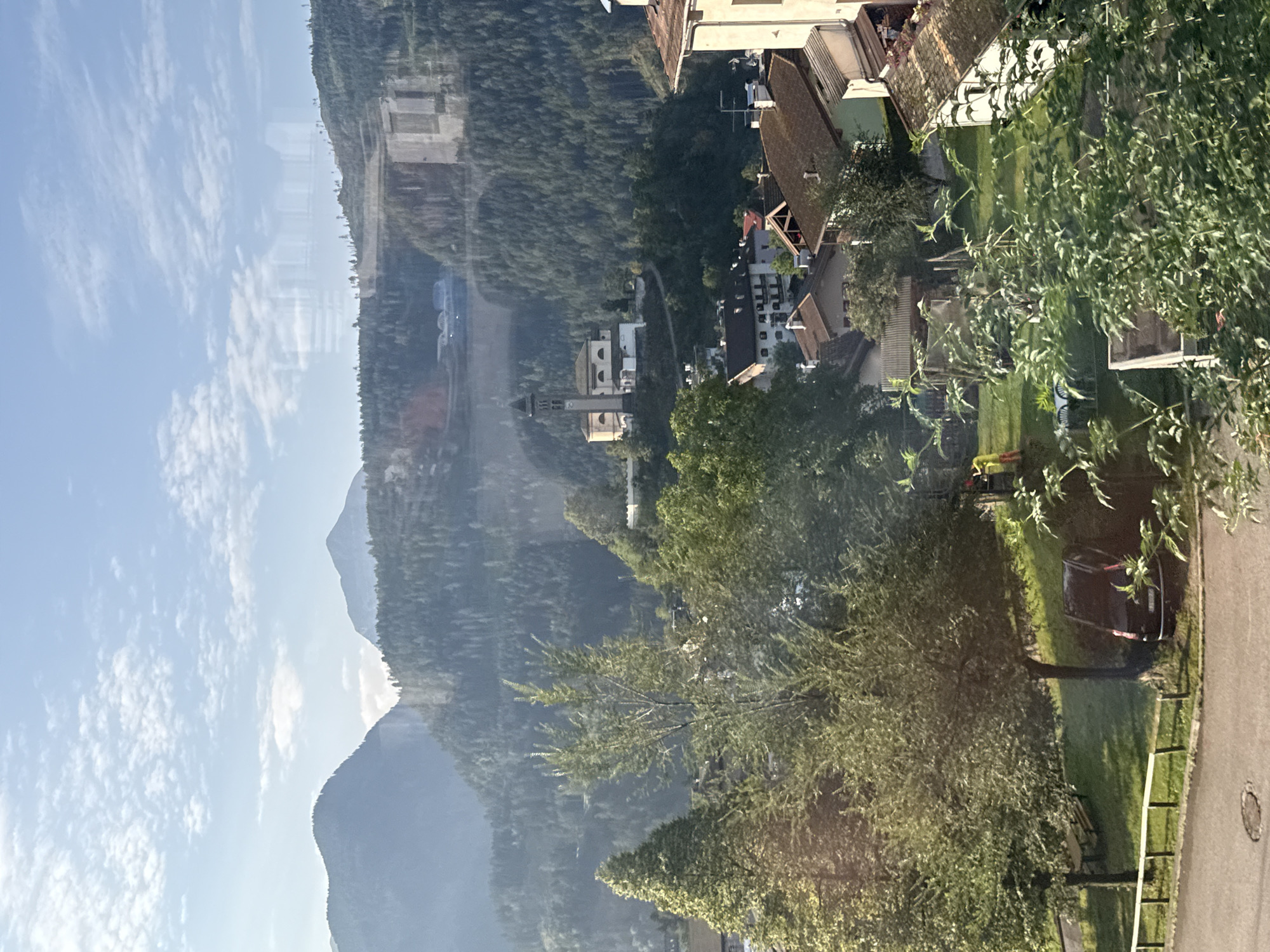 The image depicts a mountainous landscape with a village nestled in the valley, likely Castello di Fiemme, Italy. In the foreground, several trees and houses are visible. A dark-colored car is parked on a grassy area next to a road. In the middle ground, a church with a bell tower stands on a hill, surrounded by buildings and lush greenery. The background features rolling hills covered in dense forest, and further in the distance, majestic mountains under a partly cloudy sky. Reflections from a window are subtly present on the image.