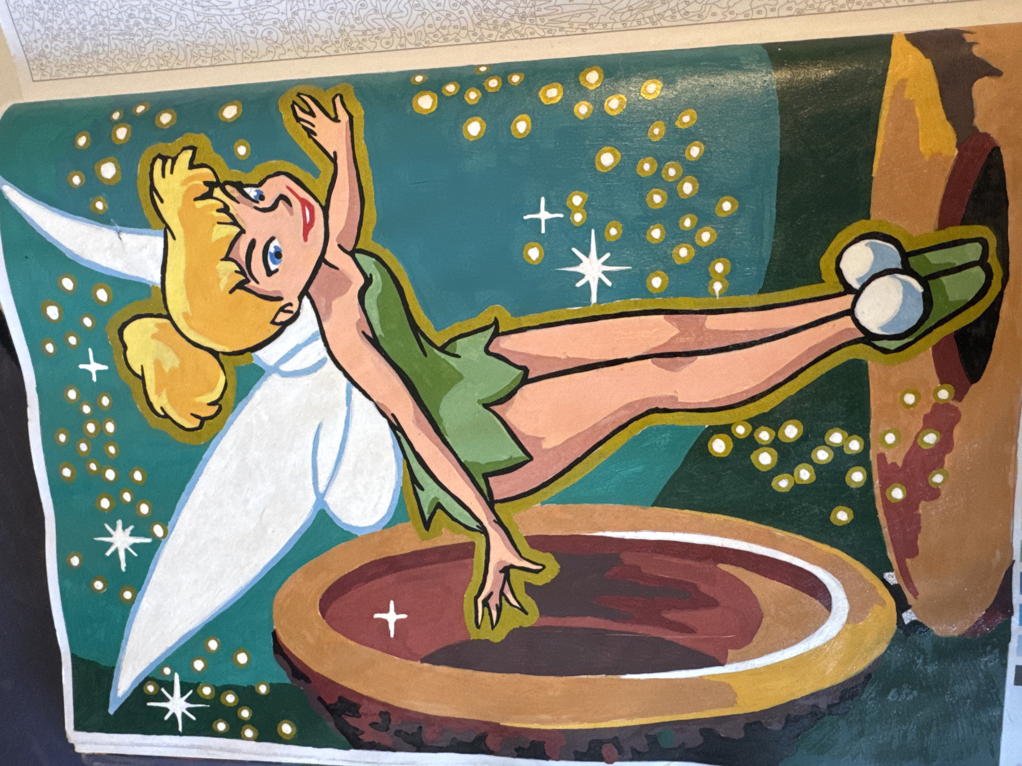 The image is a colorful painting of Tinkerbell, the Disney character, set against a teal background. She has blonde hair pulled into a bun, blue eyes, and is wearing her signature green dress. Her white wings are spread, and she is in a dynamic pose, reaching out with her left hand. To her left, there is a large, oval-shaped object with brown and red tones, resembling a portal or a keyhole. The background is adorned with yellow dots and white star-like shapes, suggesting a magical or starry environment. Tinkerbell's feet are planted on a brown surface that appears to be a platform or a part of the landscape. The painting is detailed and vibrant, with a thick black outline around the main subject and the portal. The right side of the image shows a portion of a page from what appears to be a paint-by-numbers book, with faint gray lines and numbers visible. The scene is set indoors, likely on a table or surface, as indicated by the lighting and the context of it being a painting. The location is not specified within the city of Unknown, Unknown.