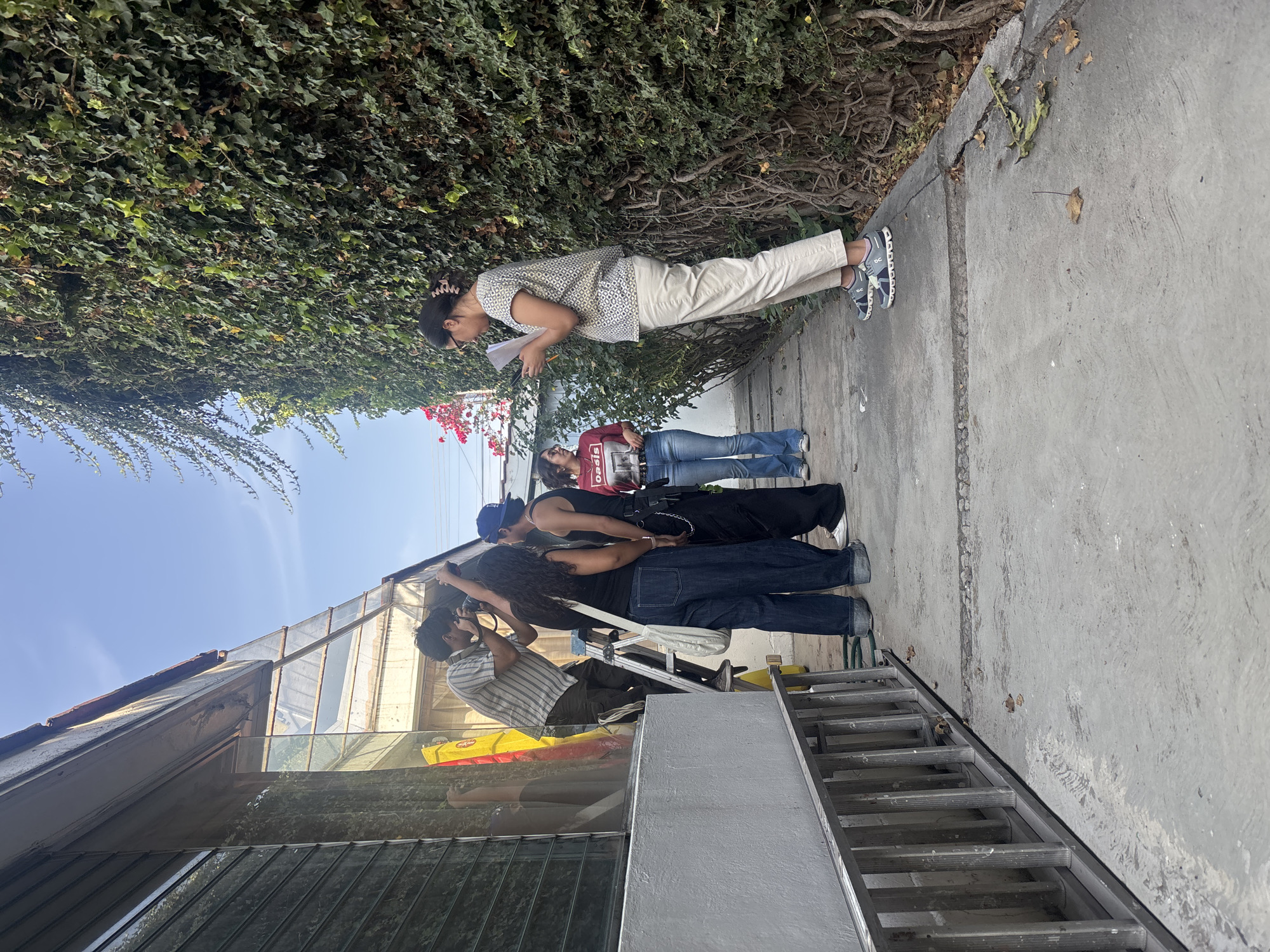 On a sunny day in Puebla, Mexico, several individuals are gathered on a concrete walkway next to a building with glass roof panels. A person standing on a ladder appears to be taking photographs, while two other people are positioned beside the ladder, seemingly involved in the photography process. Nearby, two women stand; one is holding papers and appears to be reading them, while the other woman looks towards the activity near the ladder. The walkway is adjacent to a wall covered in green vines, with hints of red flowers. A ladder is visible in the foreground, leaning against the building.