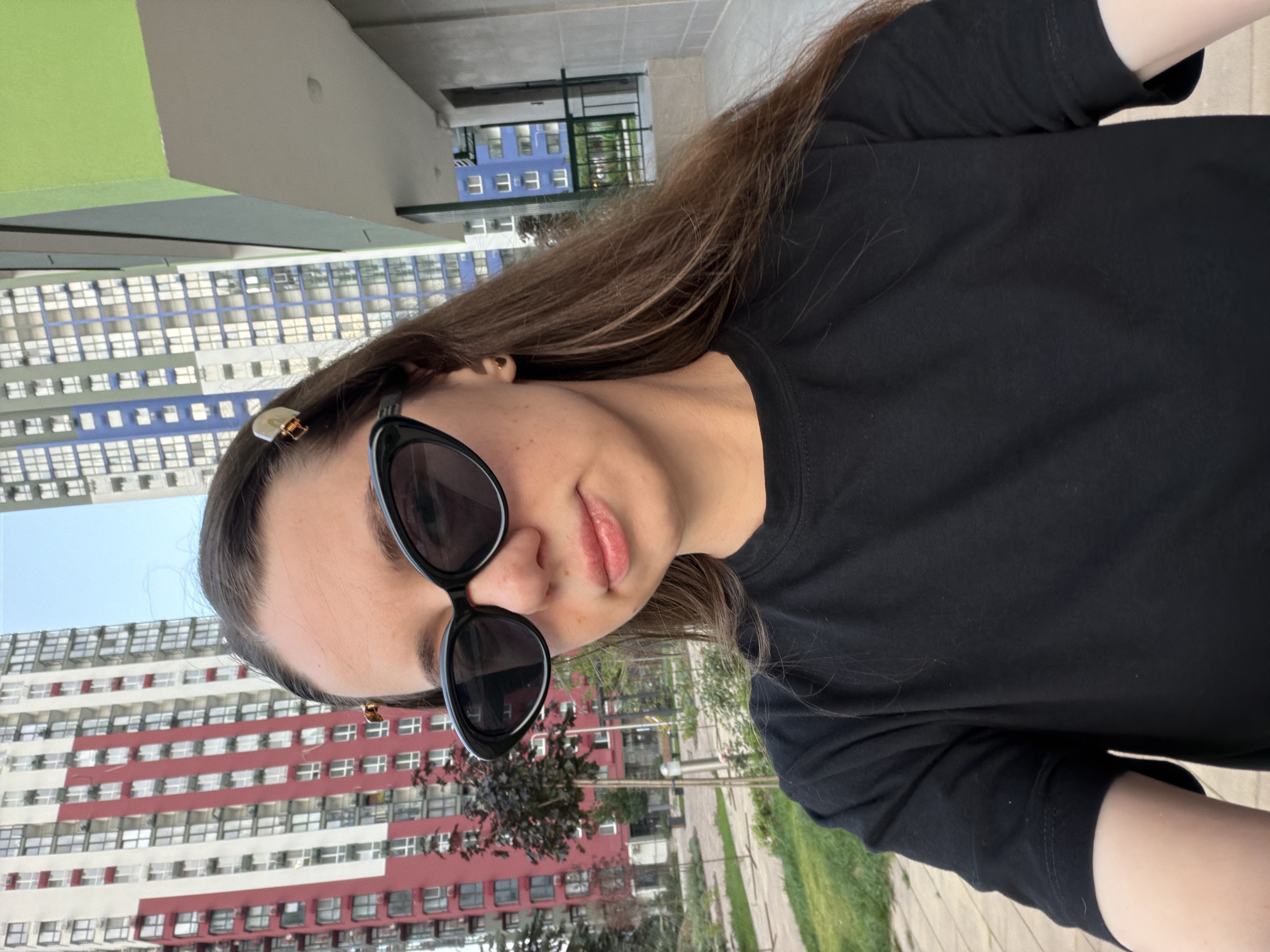 A young woman with long brown hair, wearing black sunglasses and a black t-shirt, takes a selfie outdoors. She is standing in front of a residential building with a red and white facade, and another building with a blue and white facade is visible to her right. The background suggests a city environment, likely Kyiv, Ukraine, with trees and a paved area in the foreground. The woman has a hair clip in her hair and a small heart-shaped earring. The lighting indicates it is daytime.