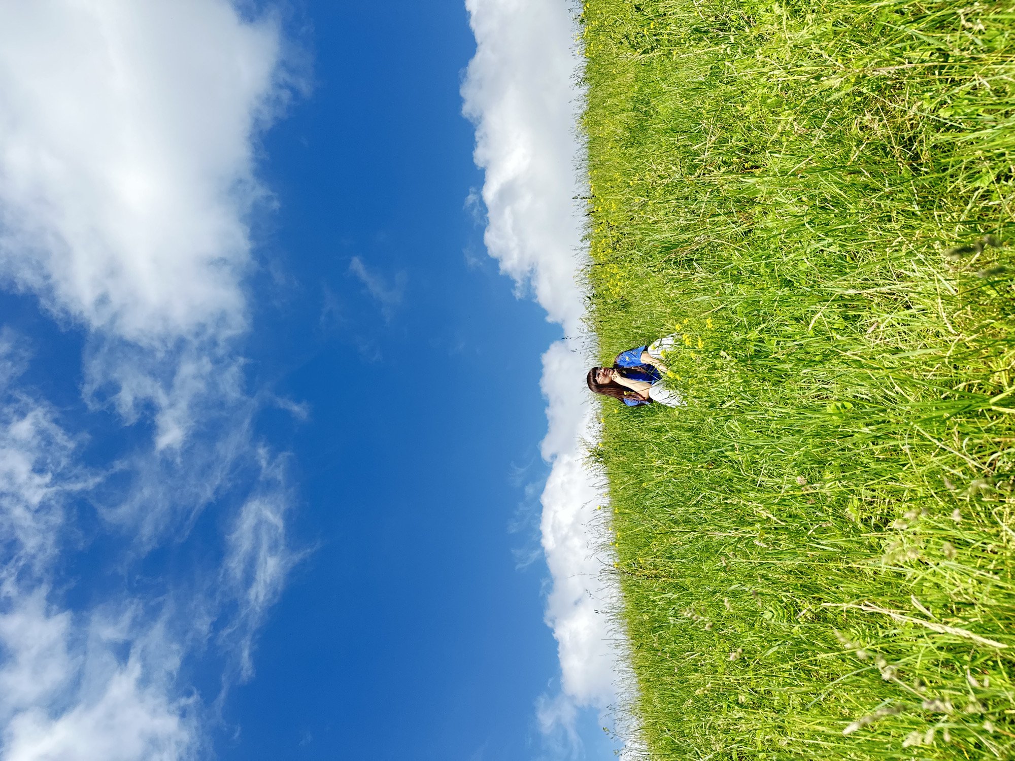 A young woman with dark hair and glasses is sitting in a field of tall green grass under a bright blue sky with scattered white clouds. She is wearing a blue shirt and white pants. The grass is dense and appears to be growing on a gentle slope. Small yellow flowers are visible interspersed within the grass. The woman is looking to her right and resting her chin on her hand, appearing contemplative. The overall scene depicts a peaceful moment in a natural, outdoor setting. The image does not contain enough information to definitively place the location in the city of Среднее Кечево, Russia.