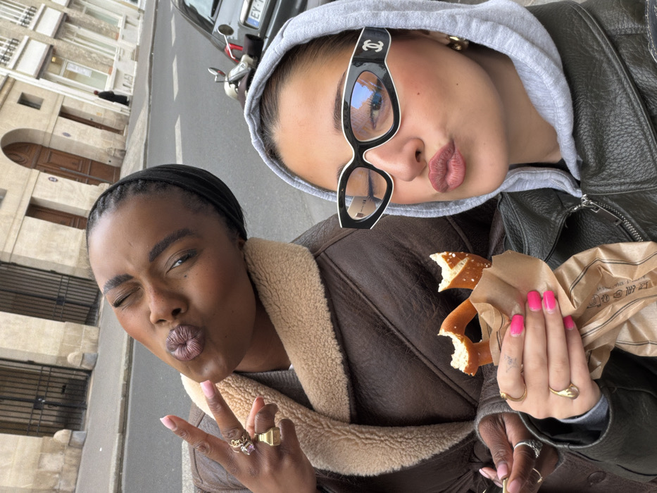 Two individuals pose for a close-up selfie on a street in Paris, France. The background reveals light-colored stone buildings with arched doorways, a parked silver car displaying a French license plate (EY-739-GF), and a scooter. The individual on the left, a Black woman, has dark skin, braided hair with a dark wrap, and wears a brown faux-leather jacket with a tan shearling collar. She winks with her right eye, puckers her lips, and makes a peace sign with her left hand, which is adorned with multiple rings and light pink nail polish. The individual on the right, a woman with a lighter complexion, wears a gray hooded sweatshirt under a dark jacket and prominent black and white Chanel glasses. She also puckers her lips and holds a partially eaten, salted pretzel in a brown paper bag with her right hand, her nails painted bright pink. Gold-toned jewelry is visible on both individuals' hands and the right individual's ear.