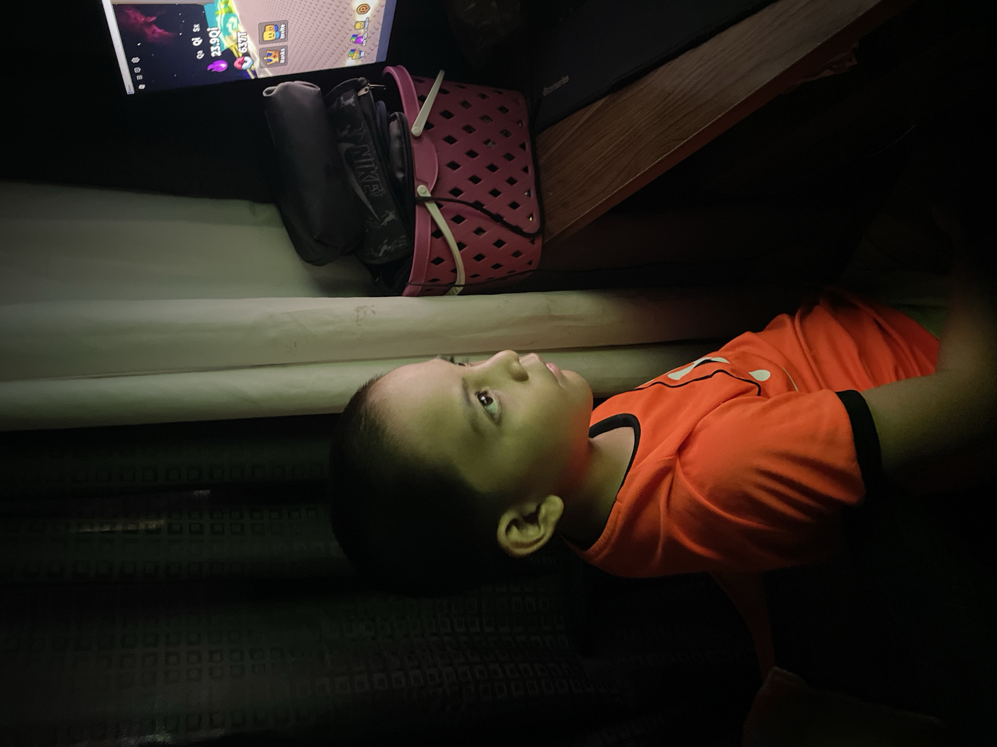 In a dimly lit room, a young boy in an orange t-shirt is captivated by a computer screen. The screen displays a game interface with colorful graphics and text, suggesting an interactive digital environment. Beside the computer, a pink woven basket holds several dark bags, possibly belonging to the boy or an adult. The room appears to be a personal space, perhaps a bedroom or study, with dark curtains and a white curtain visible in the background. The boy's posture and focused gaze indicate deep engagement with the game. While the image does not explicitly provide information about the location within Davao City, Philippines, the presence of common household items suggests an indoor setting.