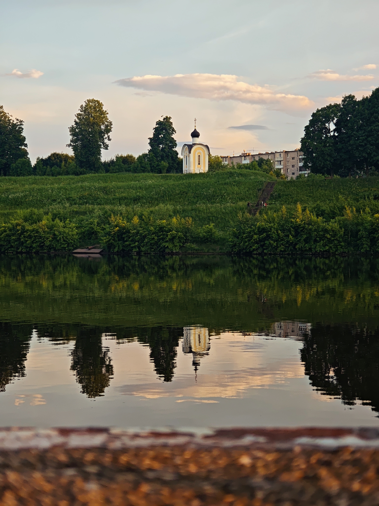 A vertical, eye-level, full shot captures a serene waterside scene at dusk in Vodnik Snt, Russia. The foreground is dominated by a blurred, reddish-brown textured surface, possibly a railing or bench, leading the eye toward the main subject. The middle ground presents a calm body of water reflecting the sky and the scenery above. On the far bank, a lush green hillside rises, dotted with trees and shrubs. Perched on the crest of the hill is a small, white and yellow chapel with a dark dome and cross. Behind the chapel, a multi-story, grey apartment building is visible. The sky above is a soft blend of blue and pale pink, with scattered clouds tinged by the setting sun. The reflection in the water mirrors the chapel, the trees, and the apartment building, creating a symmetrical and tranquil composition. No people are clearly discernible in the scene.