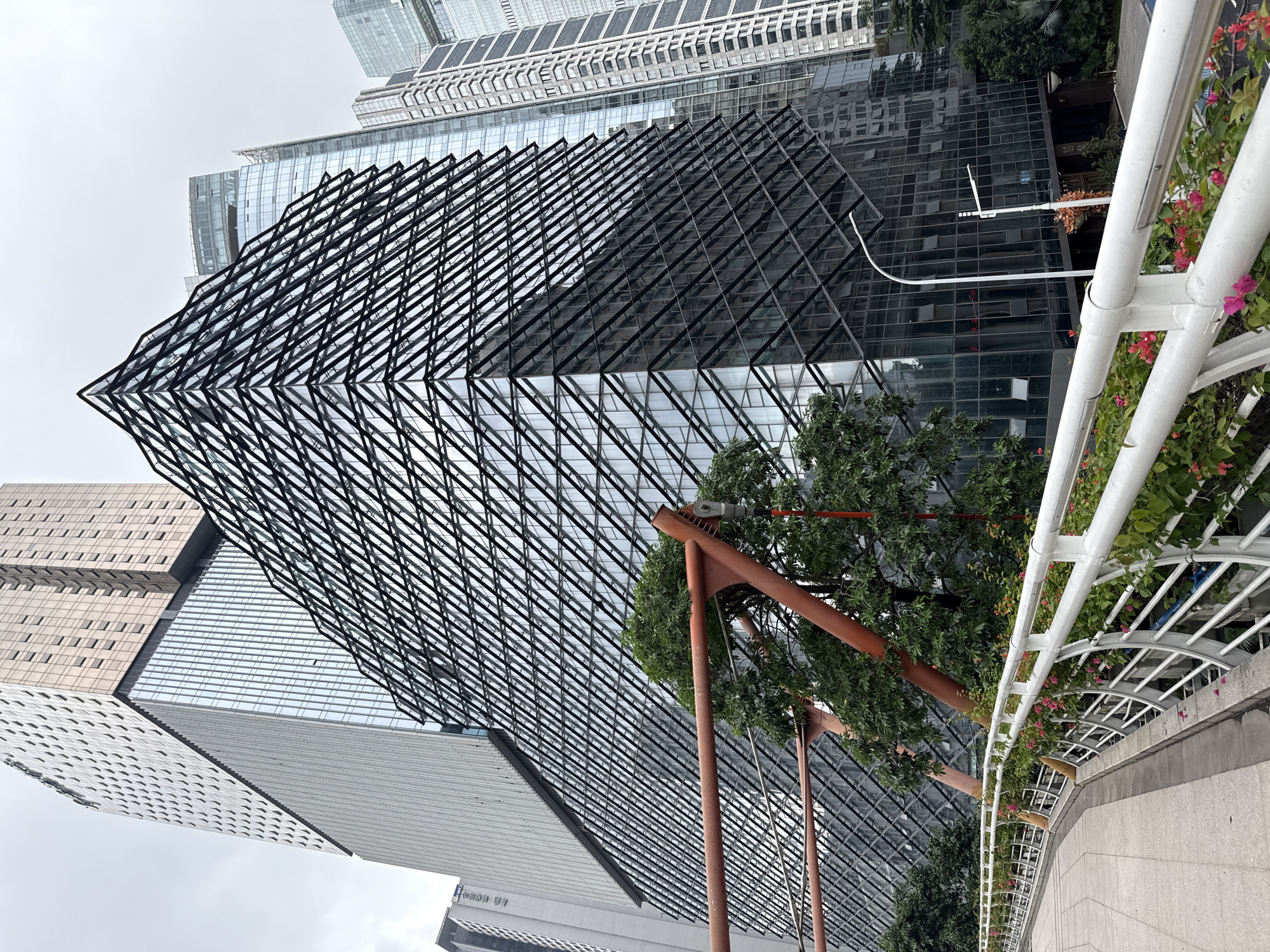 The image shows a modern architectural scene in Tianhe District, China, under an overcast sky. In the foreground, a curved walkway with a white railing is lined with flowering plants. Beyond the walkway, an orange-colored structural element, possibly part of a bridge or overpass, extends horizontally and diagonally, with green trees visible behind it. The central focus of the image is a striking building with a glass facade featuring a distinctive, angular, diamond-patterned external structure. Adjacent to this building are several other tall, contemporary skyscrapers with glass exteriors, reflecting the muted sky and surrounding architecture. The scene is devoid of people and activity, presenting a static view of urban design and infrastructure.