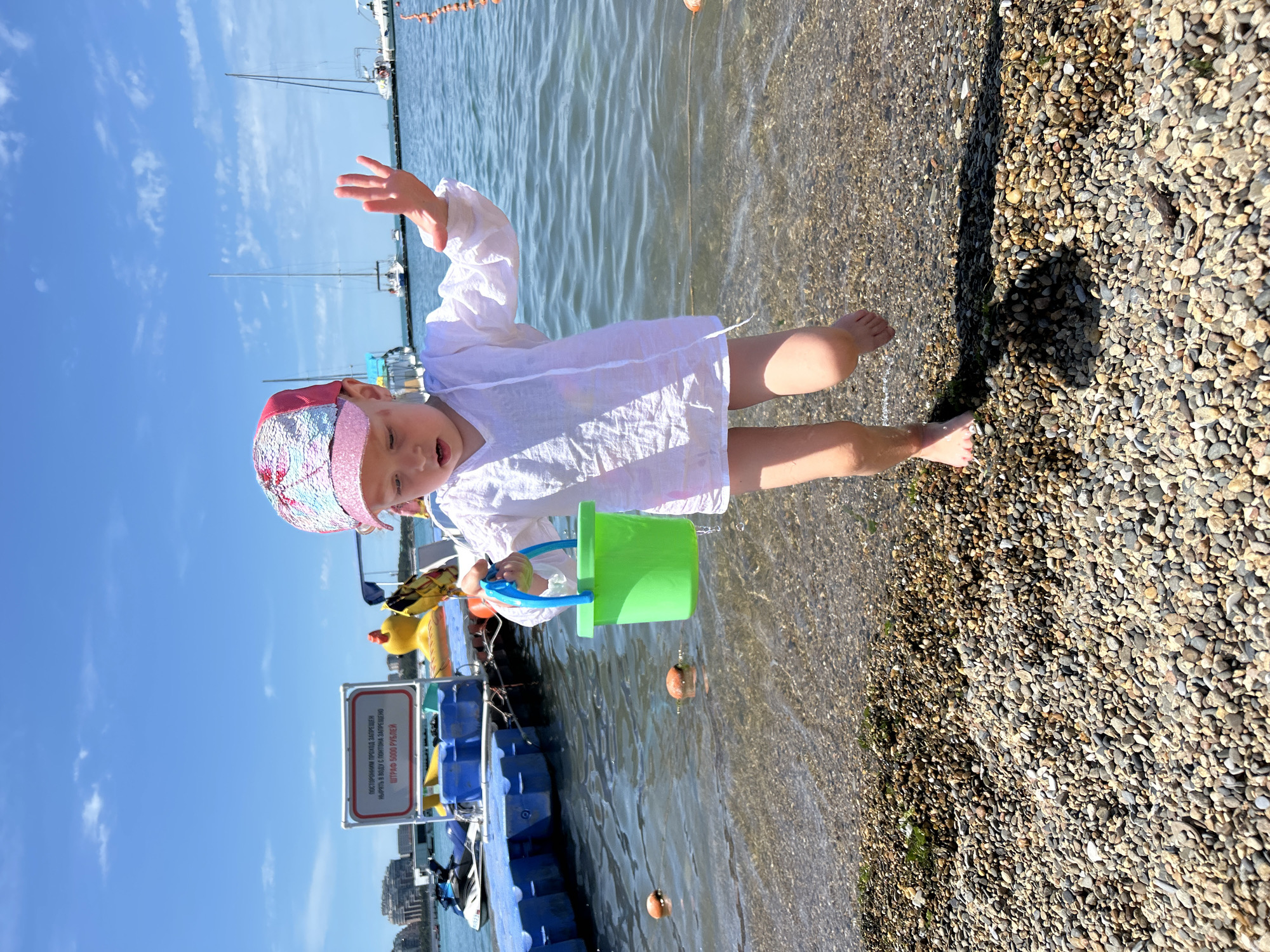 A young Caucasian child wearing a white cover-up and a colorful sequined baseball cap stands on a pebble beach next to the water. The child holds a bright green plastic bucket with a blue handle. The child's left arm is raised, and their right leg is extended forward as if stepping into the water. In the background, there are several boats docked in the sea, and a city skyline is visible in the distance. The sky is blue with scattered white clouds. The scene is located on the Gelendzhik Bay waterfront in Gelendzhik, Russia.