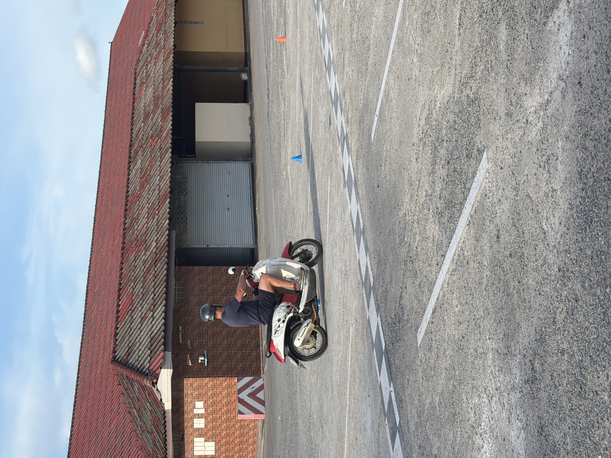 In Baan Mae Nam, Thailand, a man wearing a dark blue t-shirt, dark shorts, and a gray helmet is riding a red and white scooter on a paved surface. The scooter has black tires and a silver exhaust pipe. The man is facing to the right and holding the handlebars. In the background, there is a building with a red tiled roof, made of red brick and yellow siding. A gray metal roll-up door is visible on the building. The ground is marked with white lines, and there are two cones, one blue and one orange, on the pavement. The sky is blue with some clouds.