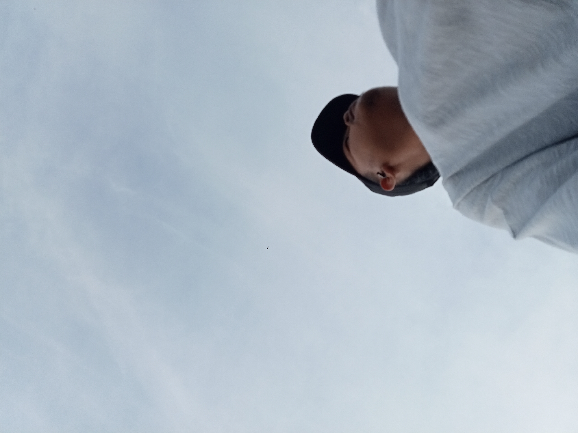 An individual wearing a dark baseball cap and a light-colored hooded sweatshirt is viewed from a low angle, looking upwards against a pale, overcast sky. The person's head is tilted back, and their face is partially visible, showing an ear and the side of their nose. The sweatshirt is made of a textured fabric and drapes over their shoulder. In the upper left portion of the sky, a small, dark silhouette of a bird is visible. The overall scene is dominated by the sky, with the individual positioned in the upper right quadrant. The image appears to be a candid shot taken outdoors. There is no identifiable information to place this scene in Kualu, Indonesia.