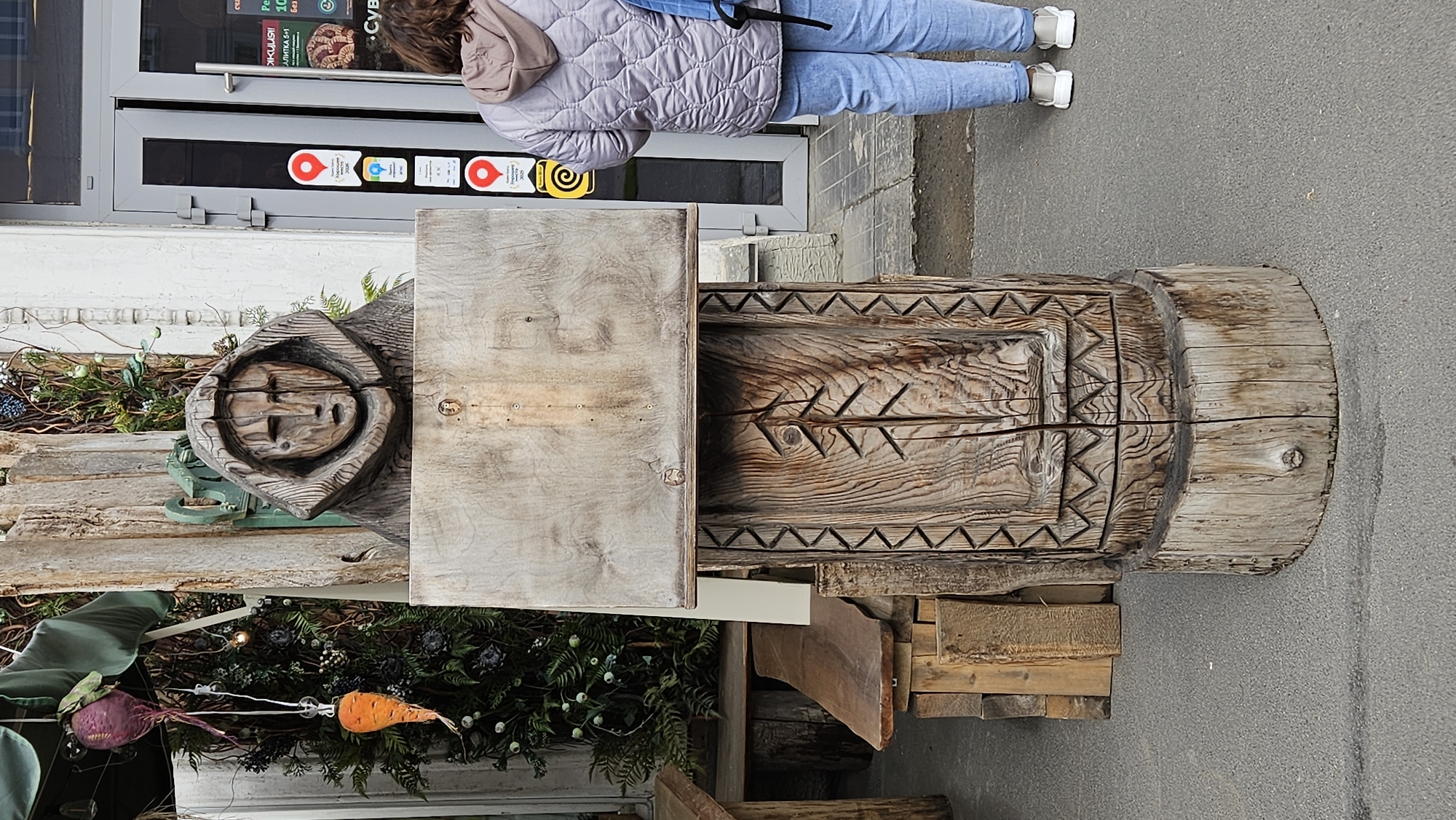 A person in jeans and a puffer jacket stands in front of a store, looking at a wooden sculpture in the city of Unknown, Unknown. The sculpture features carved faces and geometric patterns. Vegetables, including a beet and a carrot, are displayed with foliage near the sculpture. The storefront has signs with logos. The surrounding environment includes the sculpture, a sidewalk, and parts of the store's exterior.