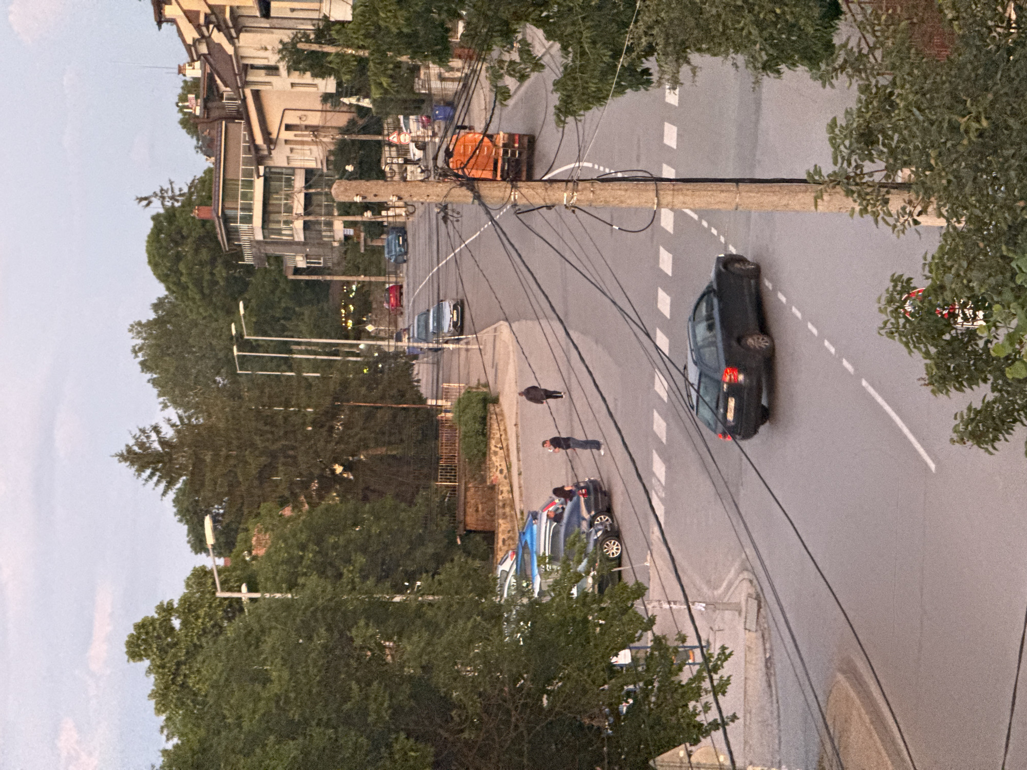 The image depicts a street scene in Sofia, Bulgaria, likely during the late afternoon or early evening, with a dusky sky. The foreground shows a dark-colored sedan driving on a wet road, with a pedestrian crossing visible. A large concrete utility pole, heavily laden with electrical wires, stands prominently near the road. In the middle ground, parked cars are visible on the left side of the street, next to a sidewalk bordered by trees. A few people are standing near the parked cars, with one person appearing to hold a phone. Further down the street, an orange truck with its back raised is moving away from the viewer. On the right side of the image, a row of multi-story buildings with balconies and tiled roofs line the street, interspersed with trees. The overall impression is that of a residential urban environment.