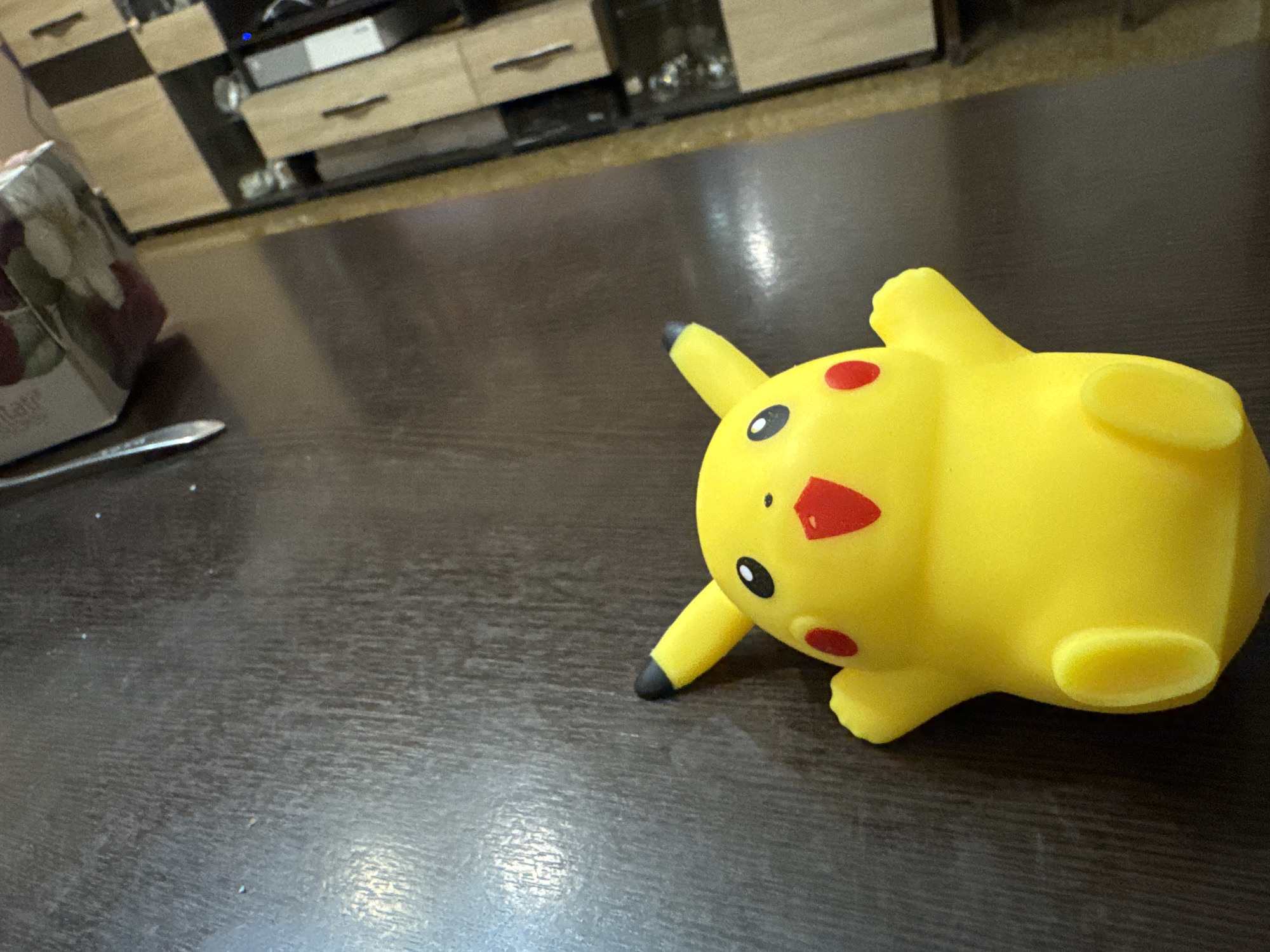 A yellow Pikachu toy lies on its side on a dark brown wooden surface. A package is to the left and behind the Pikachu. The background includes a light brown wooden cabinet and black electronic equipment. This scene is not identifiable as being in Yerevan, Armenia.