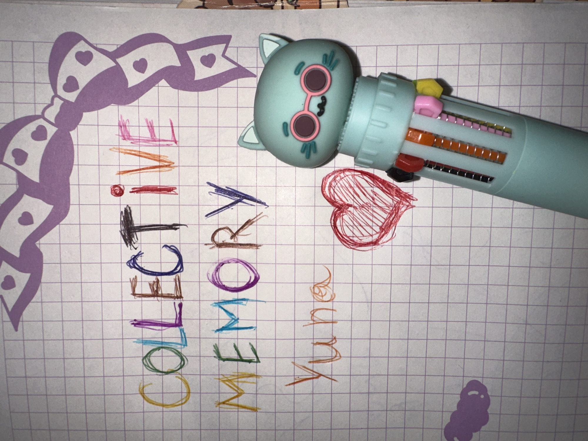The image shows a notebook with a grid pattern on its pages. Written on the page in colored ink is the phrase "collective memory Yuma". There is a cartoon cat-shaped multi-color pen resting on the page, next to a red heart doodle. To the left, there's a purple drawing of a bow with heart details. At the bottom of the page is a doodle of what looks like a caterpillar. There is no location information given.