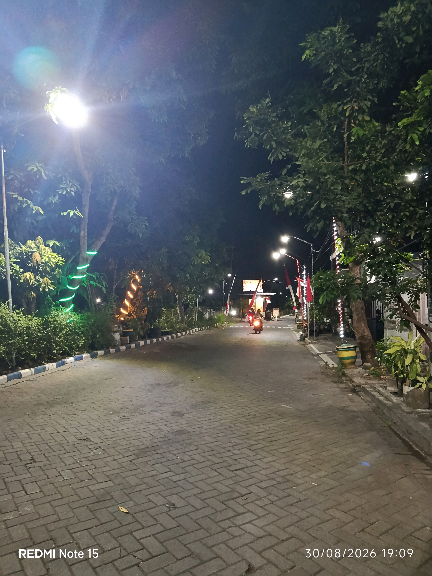 The image depicts a dimly lit outdoor scene at night, likely in Keboananom, Indonesia, as indicated by the timestamp which includes the date and time "30/08/2026 19:09". The foreground is dominated by a paved walkway made of interlocking bricks, with a slight incline. Along the left side of the walkway, there is a curb with alternating blue and white patterned blocks. Lush green foliage and trees line the walkway, some of which are decorated with festive lights. Specifically, one tree on the left has bright green lights spiraling around its trunk, while another tree further down the path has orange lights wrapped around it.
Several streetlights illuminate the area, casting a yellowish glow. In the mid-ground, a person is riding a scooter with its headlights on, moving away from the camera. The scooter appears to have a red top. Further in the distance, more trees and what appear to be buildings or structures are visible, also lit. Red and white flags, possibly national flags, are mounted on poles along the right side of the walkway, near the trees and a trash bin. The overall atmosphere is that of a public space or park at night, decorated for an event or general ambiance. The bottom left corner of the image displays the text "REDMI Note 15", indicating the device used to capture the photograph.