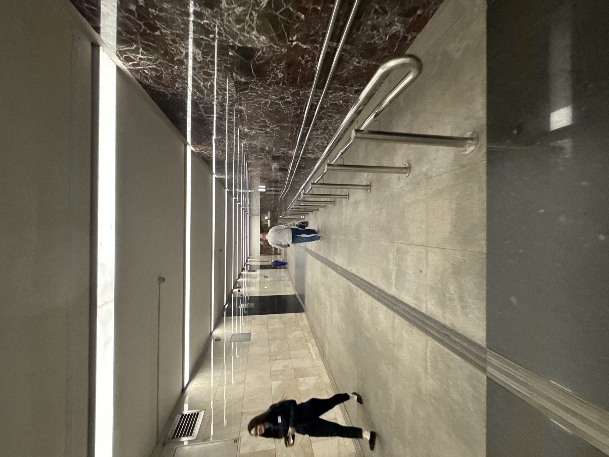 The image shows a wide, brightly lit pedestrian tunnel with polished stone walls and a tiled floor. The tunnel appears to be underground, possibly in a subway station or a pedestrian underpass. A ramp with a metal handrail runs along the right side of the tunnel. In the foreground, a woman with dark hair wearing a dark uniform and pants is walking to the left, holding a small black object. Further down the tunnel, a man in a light-colored jacket and blue jeans is walking away from the camera, carrying a dark bag. A small child in a blue outfit is visible in the distance, walking towards the man. The lighting is provided by long fluorescent fixtures on the ceiling. The walls on the left are clad in light-colored marble, while the wall on the right features dark red marble with white veining. A tactile paving strip is visible on the floor running down the center of the tunnel. This location is the pedestrian underpass at the metro station "Prospekt Mira" of the Kaluzhsko-Rizhskaya line in Moscow, Russia.