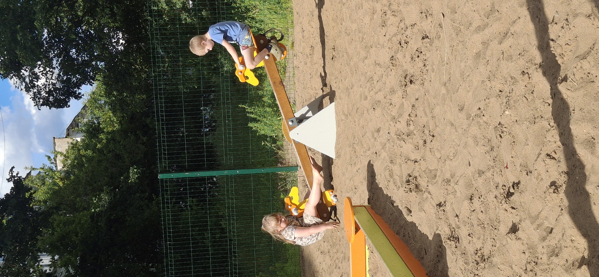 In a sandy playground, a seesaw is in motion. On the higher end, a young boy with blond hair, wearing a blue t-shirt and patterned shorts, is seated, leaning forward. His legs are bent, and he appears to be enjoying the ride. On the lower end of the seesaw, a young girl with long blond hair, dressed in a light-colored floral dress, is positioned, her legs extended upwards. She is also seated, and her expression suggests she is engaged in the playful activity.
The seesaw itself is made of wood with yellow animal-shaped seats. It is supported by a white triangular structure. The playground is covered in sand, with visible footprints. A green metal fence encloses the area, and beyond it, lush green trees and a building are visible. The sky above is partly cloudy. The sunlight casts shadows on the sand, indicating it is daytime. The scene captures a moment of childhood fun and outdoor play. No visible text is present in the image.