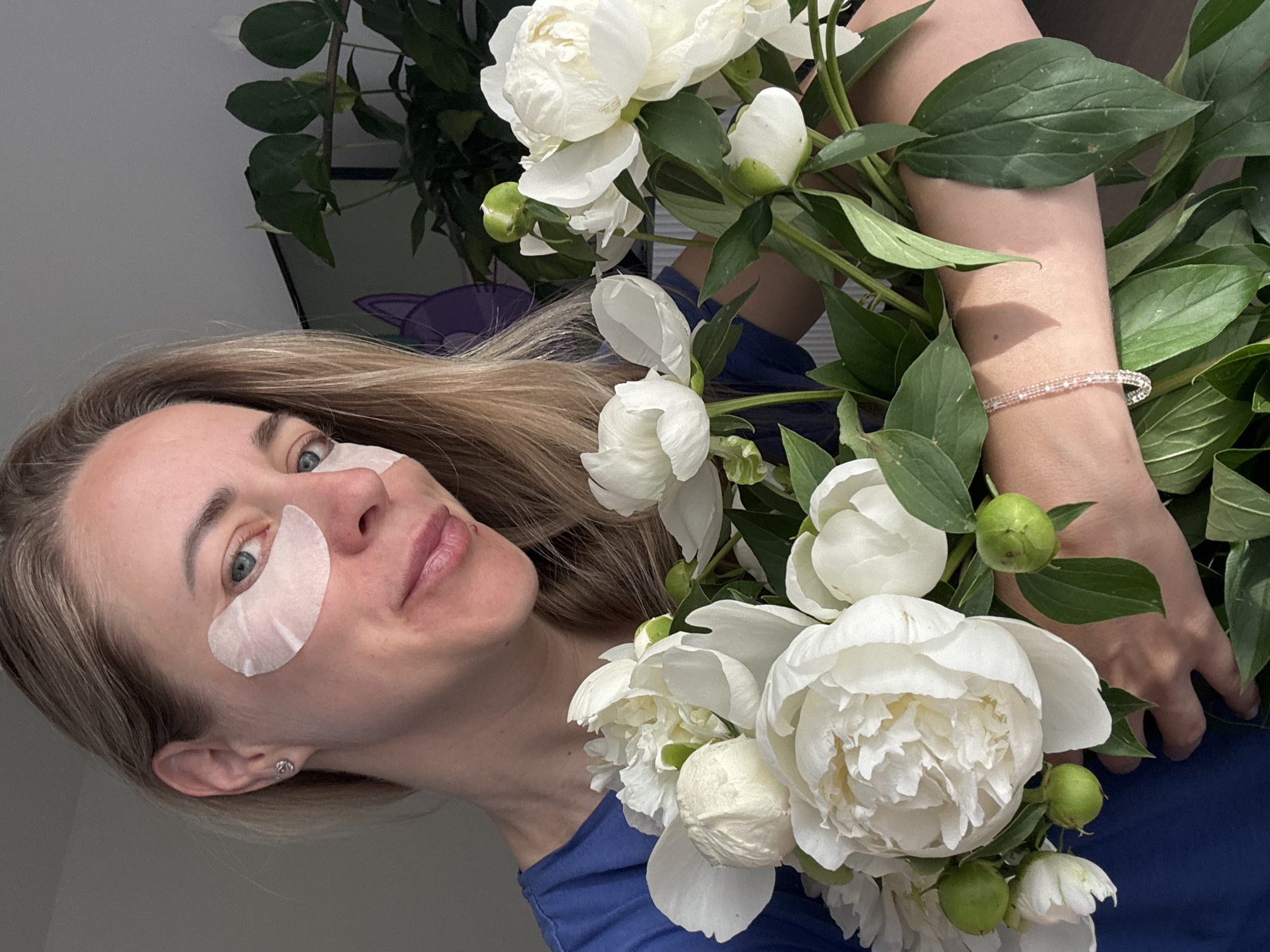A close-up, head-and-shoulders portrait of a blonde woman with blue eyes and light skin. She is wearing a blue t-shirt and has white under-eye patches on. She is holding a bouquet of white peonies and a few green buds. The background is blurred, but appears to be an indoor setting with a plant and a television screen. The woman is looking at the camera and smiling faintly. The image does not provide any information about the city of Chaikovsky, Russia.