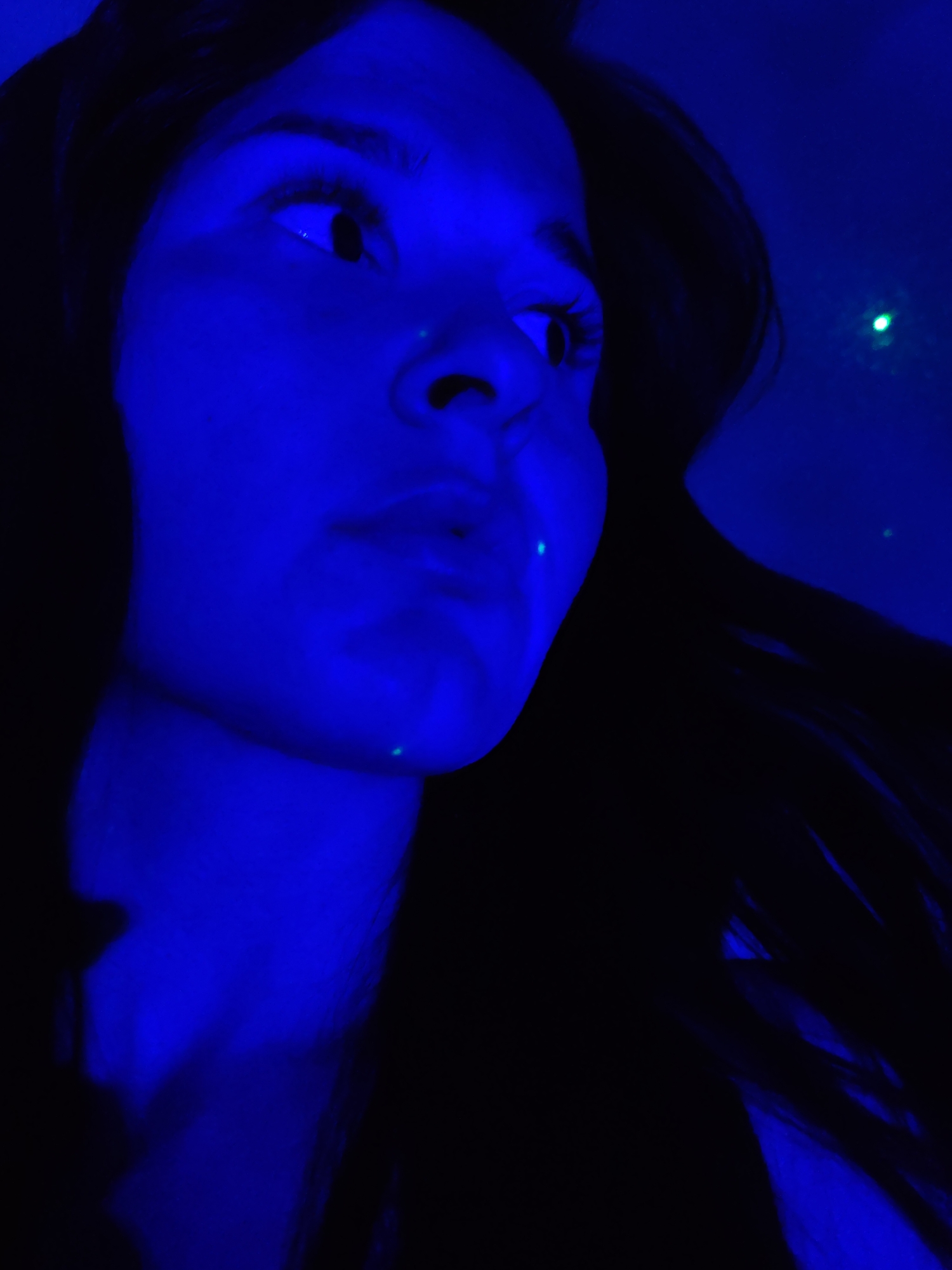 A young person with long, dark hair is bathed in intense blue light. The individual is looking up and to the right, with their face tilted slightly. A single green dot of light is visible in the background and two small green dots are present on their face, one on the nose and one on the chin. The city of Фоки, Russia, is not discernible from the image.