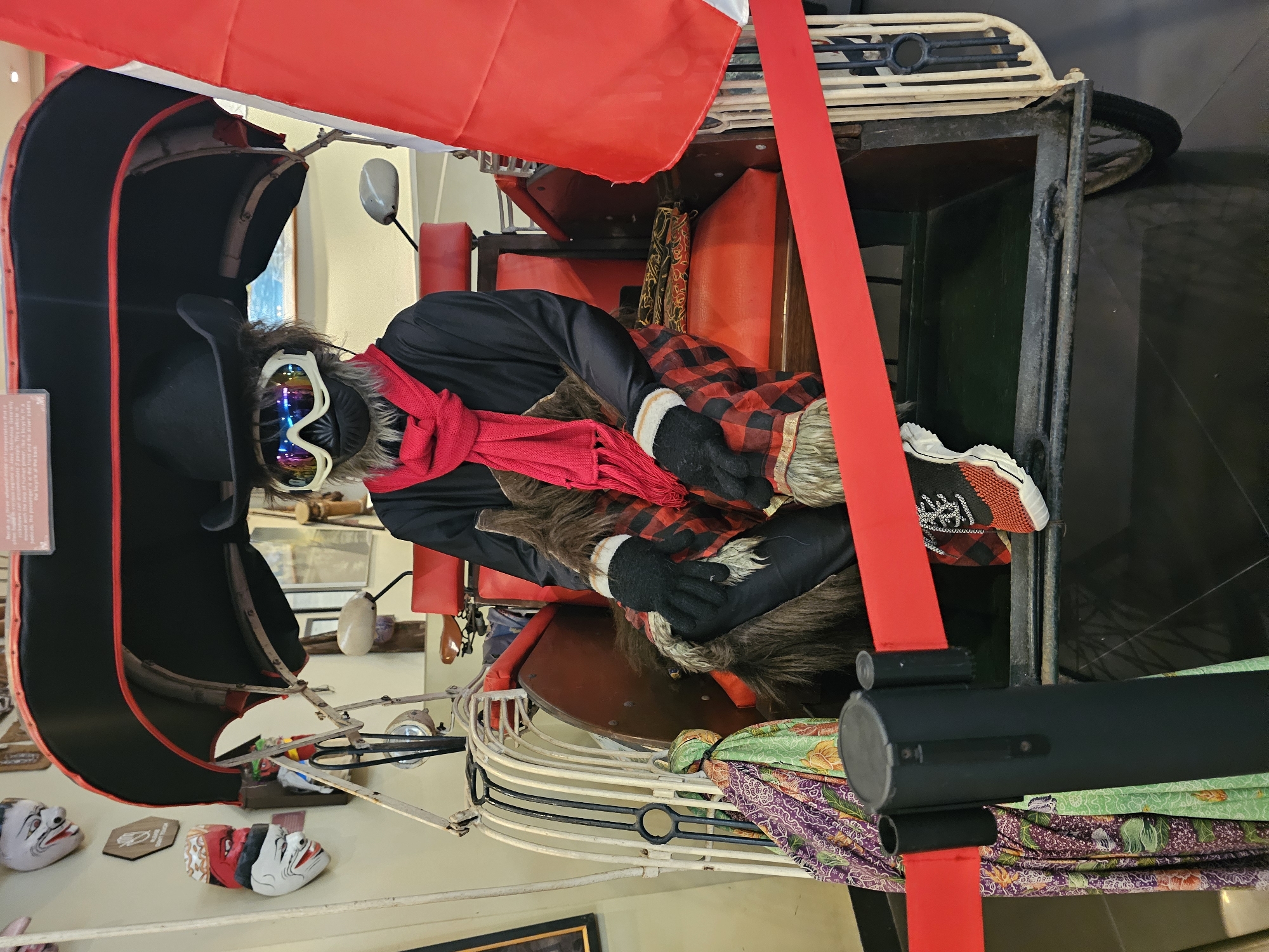 The image shows a mannequin dressed in a black jacket, red scarf, and checkered pants inside what appears to be a decorated rickshaw or similar vehicle. The mannequin has goggles and furry accents, giving it a whimsical appearance. The vehicle has a red canopy and decorative elements.
In the background, there is a shelf with masks and other decorative items. The location is identified as Kuta Selatan, Indonesia.