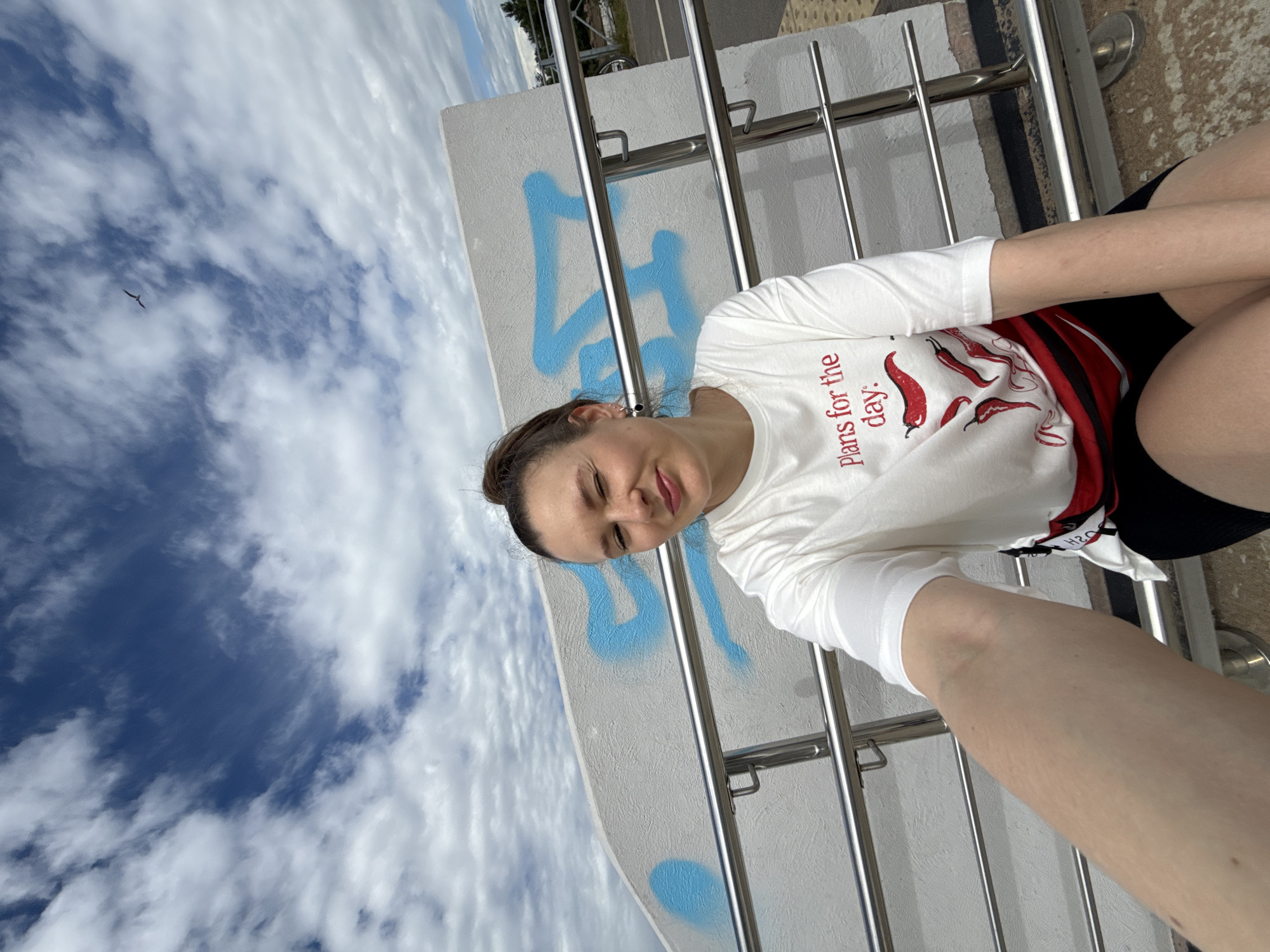 In this selfie, a woman is outdoors with a bright blue sky filled with scattered white clouds. A bird is visible flying in the distance against the sky. The woman is wearing a white t-shirt with red chili peppers and text that reads "Plans for the day.". She is sitting next to a metal railing and a white wall with blue spray paint graffiti. The image is taken from a low angle, looking up towards the sky. It is daytime, and the weather appears to be pleasant. The woman has her eyes closed and a slight smile, suggesting a relaxed or happy mood. The scene appears to be in an urban or park setting, possibly a promenade or an elevated walkway, given the railing and wall. The presence of graffiti and the style of the railing suggest a modern, possibly recreational area.