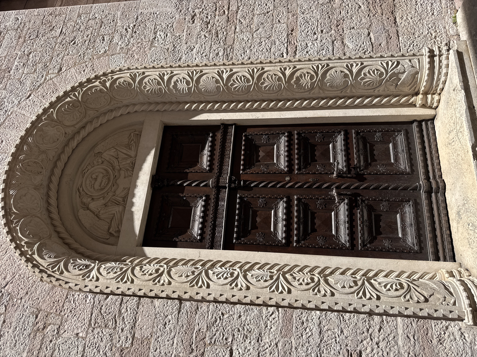 The image shows a close-up of a richly carved wooden double door set within a stone arched doorway. The door is dark brown and features intricate paneling with square, raised sections bordered by detailed carvings. The archway itself is made of light-colored stone and is adorned with elaborate decorative moldings. The outer edge of the arch features a zigzag pattern, while the inner edge has a rope-like twist. Beneath the apex of the arch, a carved relief depicts a scene with figures, including what appears to be an angel kneeling. The surrounding wall is constructed of rough-hewn, pinkish stone blocks. The scene is illuminated by bright sunlight, casting shadows that accentuate the carvings.