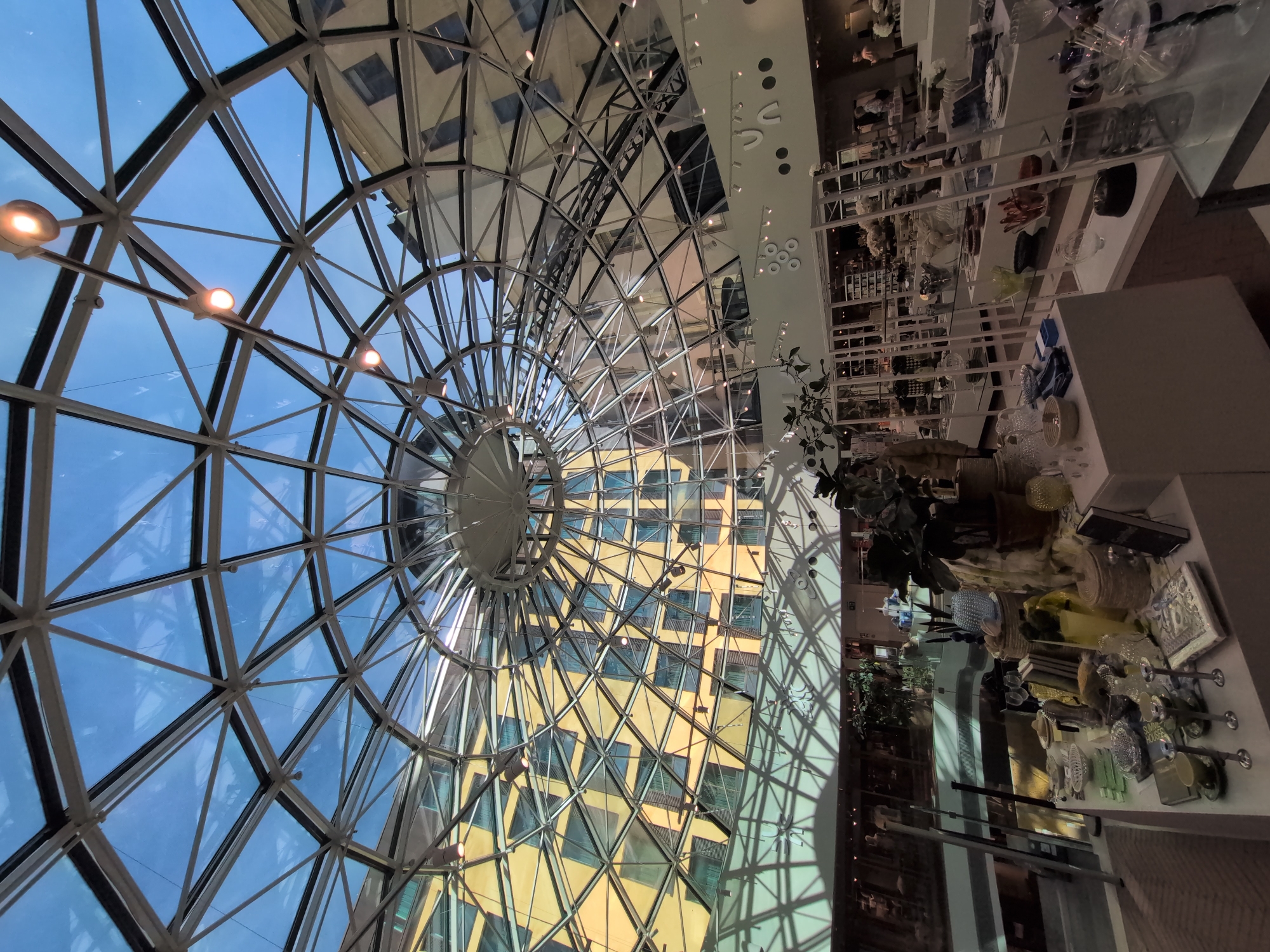 The image shows the interior of a modern retail space with a geodesic glass dome ceiling. The dome is a prominent feature, allowing natural light to illuminate the area. The reflection of a yellow building with a grid-like facade is visible on the glass dome. Inside the retail space, there are display shelves filled with various items, likely home decor or gifts, including glassware, books, and decorative objects. Several potted plants are also present, adding greenery to the environment. A few individuals are visible browsing or working in the store, though their features are not distinct. The overall atmosphere is bright and contemporary. This image was taken in the Stockmann department store in Helsinki, Finland.