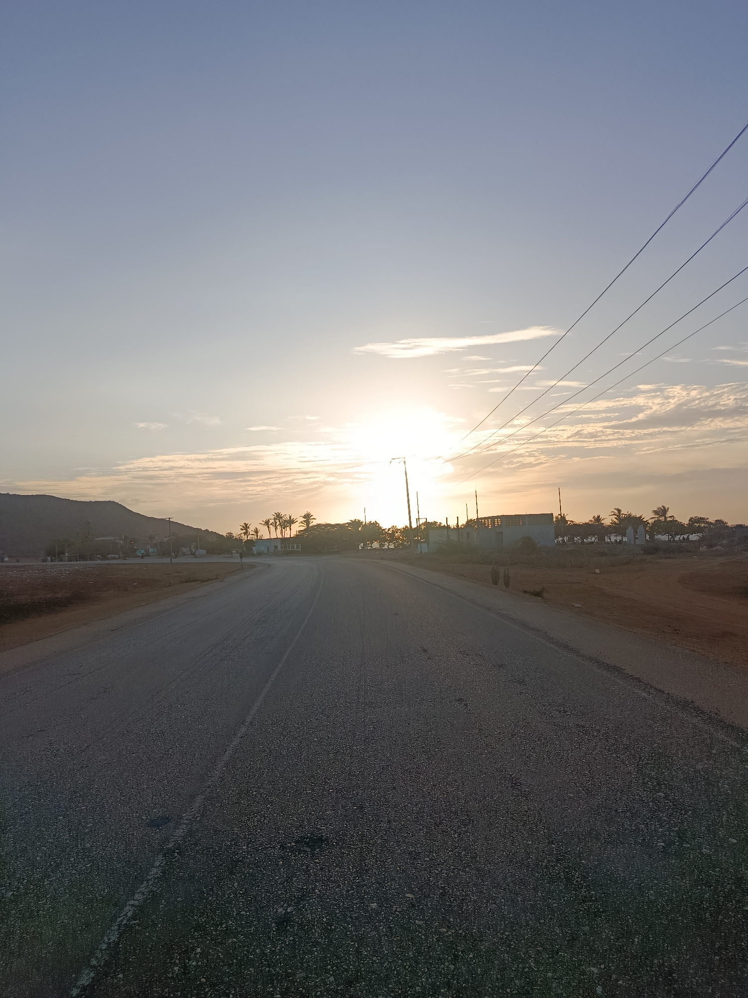 The image depicts a paved road in Altagracia, Venezuela, leading toward a bright sun near the horizon, partially obscured by thin clouds. Utility poles with visible power lines are located to the right of the road. Buildings and vegetation, including palm trees, are visible on the horizon. A low mountain is present on the left side of the image. The road shows minor wear with broken painted lane markers. Adjacent to the road are areas of unpaved land.