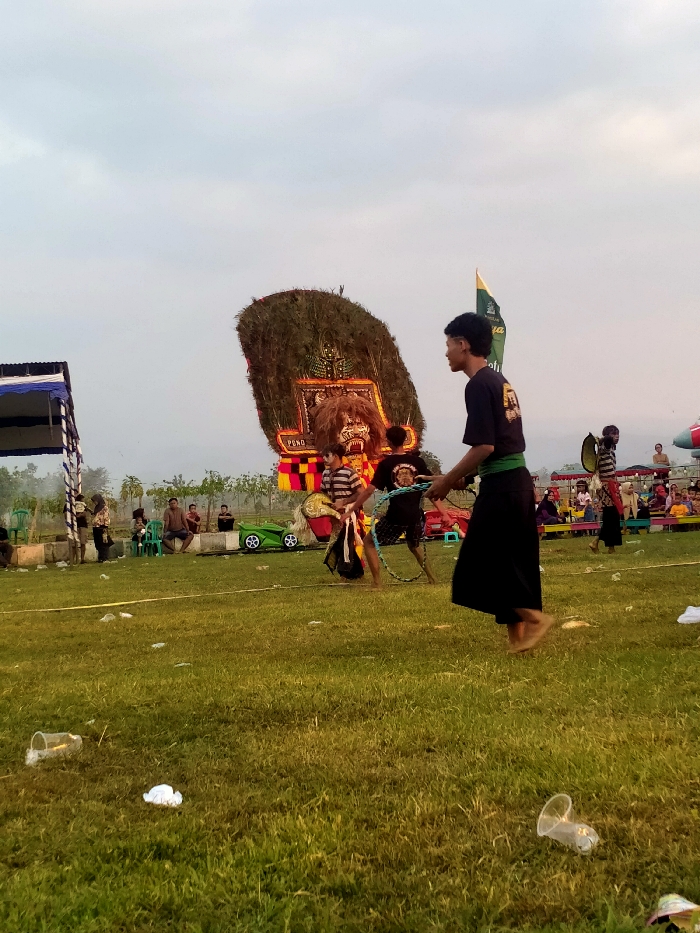 The scene depicts an outdoor cultural performance in Glonggong, Indonesia, occurring on a green grassy field under an overcast sky. In the foreground, multiple discarded clear plastic cups and white wrappers litter the ground. A barefoot male individual, wearing a black t-shirt with a yellow graphic and dark pants with a green sash, walks from right to left in the immediate foreground.
In the mid-ground, a large, elaborate traditional Indonesian "Dadak Merak" mask, adorned with green peacock feathers, a tiger-like face, and the text "PONO," is centrally positioned, likely being manipulated by a performer. To the immediate right of the "Dadak Merak" mask, another male performer in a black t-shirt and dark pants holds and manipulates a large, circular rope or chain. To the left of the "Dadak Merak" mask, a third male performer, wearing a striped shirt, holds and maneuvers a flat, horse-shaped prop. These three individuals are actively engaged in performance.
In the background, a blue and white striped canopy tent is visible on the far left. Various spectators are seated on green plastic chairs and directly on the ground, observing the performance. Some spectators are situated under additional canopies. A green stylized toy car is present near a group of seated spectators on the left. A tall, green flag with a dark emblem flies on the right side of the field. Distant trees and structures are visible on the horizon.