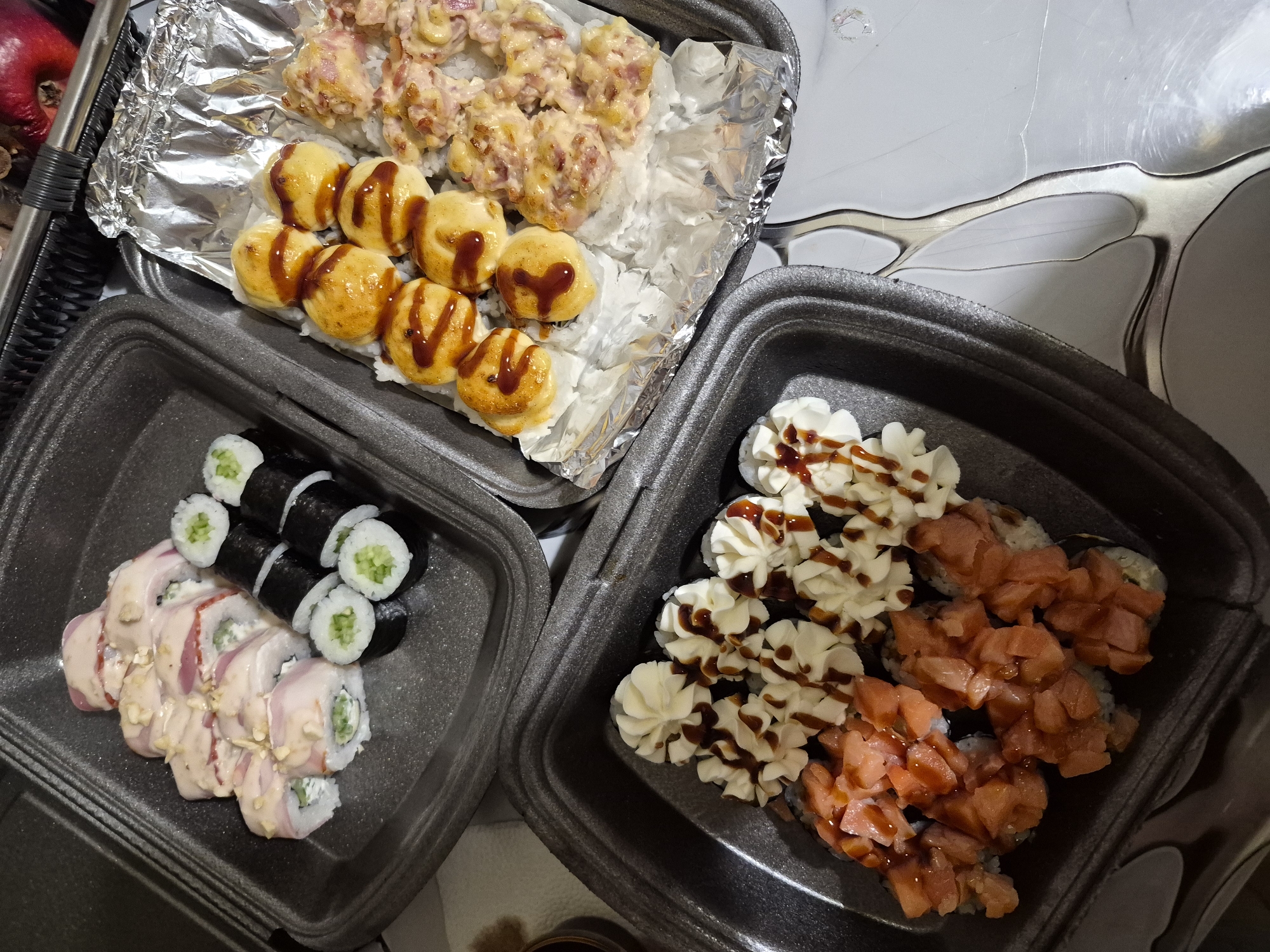 The image depicts a collection of sushi rolls in three separate containers. Two containers appear to be black styrofoam to-go containers, while the third container is aluminum foil placed on top of a black styrofoam container. The container on the left contains a mix of plain cucumber maki rolls and larger rolls topped with a light pink sauce. The container on the right contains a mix of sushi rolls topped with white cream and brown sauce, and sushi rolls topped with a salmon mixture and brown sauce. The aluminum container has two types of sushi, one with a yellow dome-like topping and brown sauce, and another with a mixture of topping which appears to be meat and cheese. The image was taken in Tobolsk, Russia.