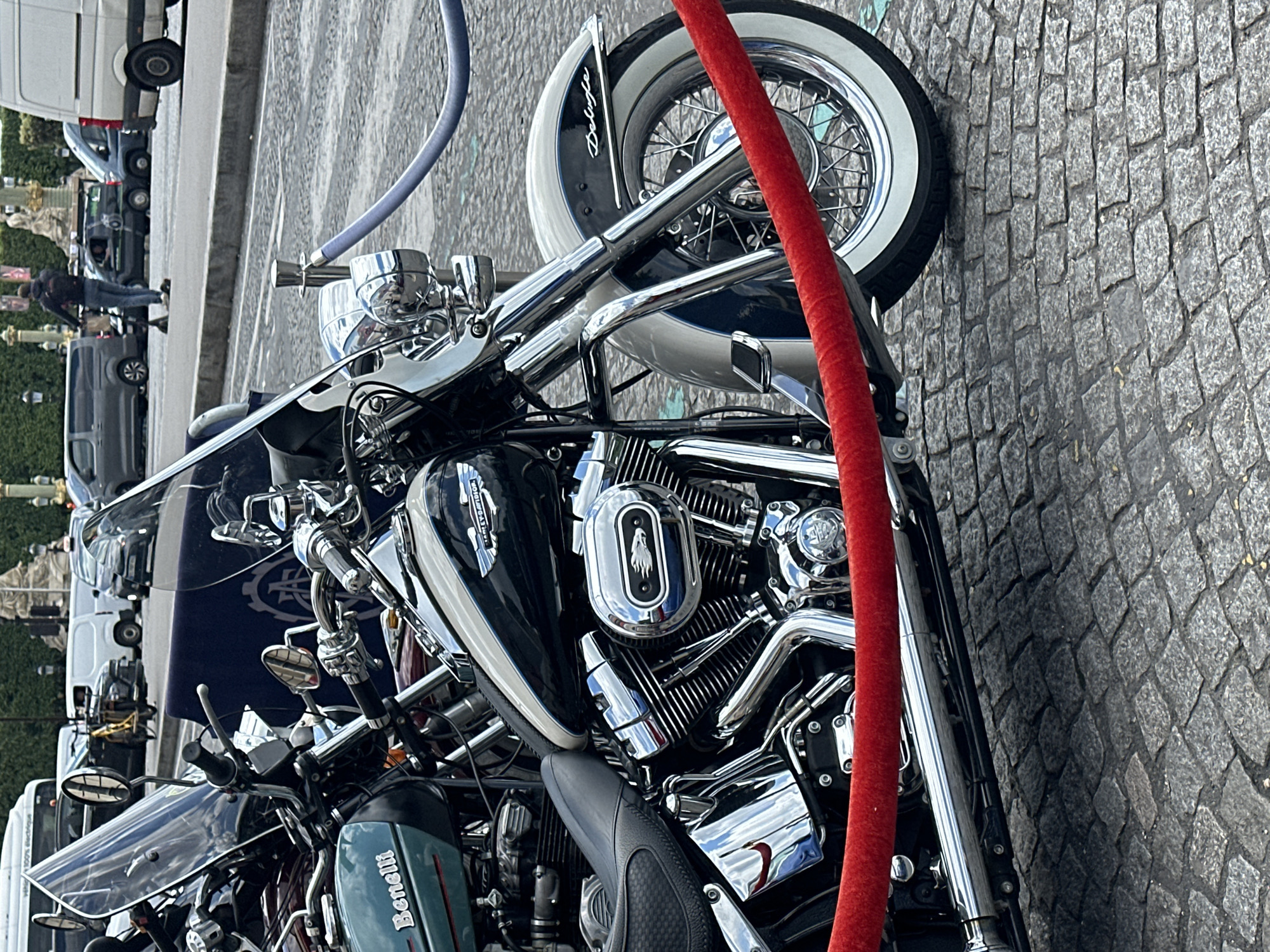 The image depicts a black and chrome motorcycle parked on a cobblestone street, partially obscured by a red velvet rope and a blue hose. The motorcycle's front tire has a wide white wall, and the engine prominently features the number "103" emblem. The motorcycle has chrome features, and a windshield. The background shows a street scene with cars and a pedestrian, and a landmark indicating the location is in Paris, France. The image is oriented sideways.