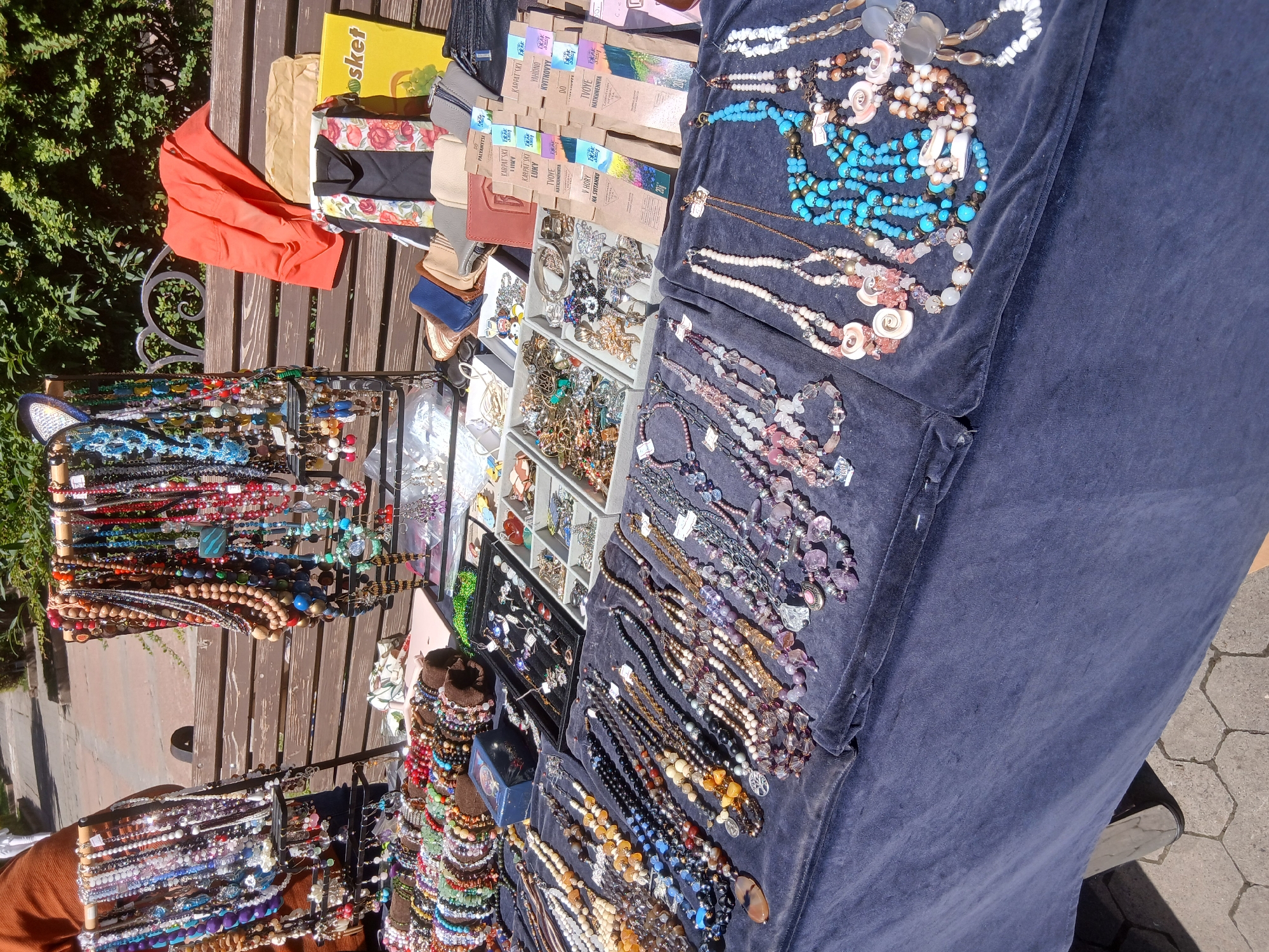 The image shows an outdoor market stall in Truskavets, Ukraine, selling a variety of handmade jewelry. The stall features a deep blue velvet tablecloth displaying numerous necklaces, bracelets, and earrings. The jewelry is made from beads, stones, and various decorative elements. In the background, a wooden bench is visible, with an orange garment and several bags or packages placed on it. Behind the bench, lush green foliage suggests a park or garden setting. The ground beneath the stall appears to be paved with hexagonal stones. The overall scene depicts a typical market day with goods laid out for display and sale.