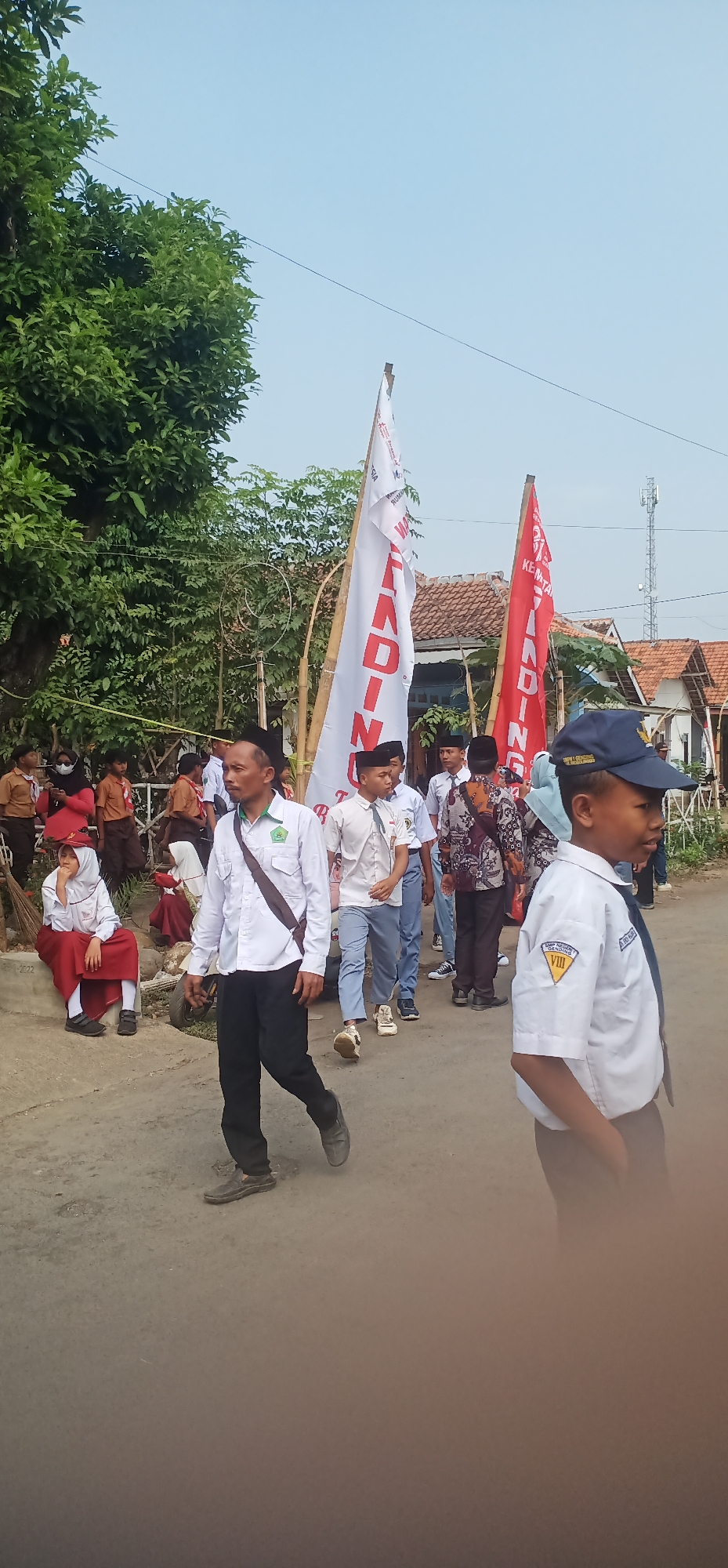 In the city of Sumberkerang, Indonesia, a group of students and adults are participating in an outdoor event. The scene unfolds on a paved road, flanked by lush green trees and modest residential buildings with red-tiled roofs. Dominating the background are two large vertical banners; one is white with red lettering, and the other is red with white lettering, both appearing to be promotional or informational.
Several individuals are visible. In the foreground, a young boy in a white uniform shirt, blue tie, and a dark blue cap with a badge is walking towards the right, his gaze directed forward. To his left, a man in a white long-sleeved shirt and black pants, wearing a dark peci (a traditional cap), walks with a brown strap across his chest. Behind him, a group of students, mostly in white shirts and dark pants or skirts, are moving along the road. Some are carrying flags or banners. On the left side of the road, two young girls in red and white school uniforms are seated on a low concrete barrier. One is looking towards the camera. The sky is clear and blue. The overall atmosphere suggests a community gathering or a school-related activity.