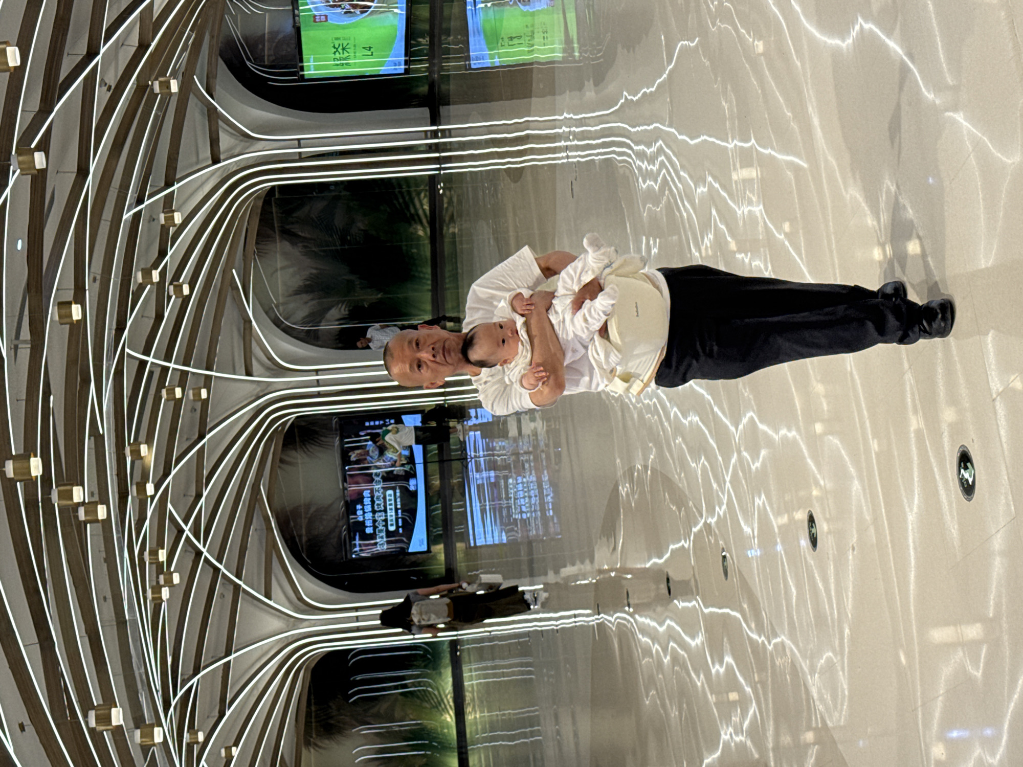 A person holds a baby in an interior location featuring curved linear structures. The location is identifiable as Liangjiang, China, based on signage in the background. The person is holding the baby in their arms. The interior space contains display screens and architectural elements. The floor is shiny and reflects light.
