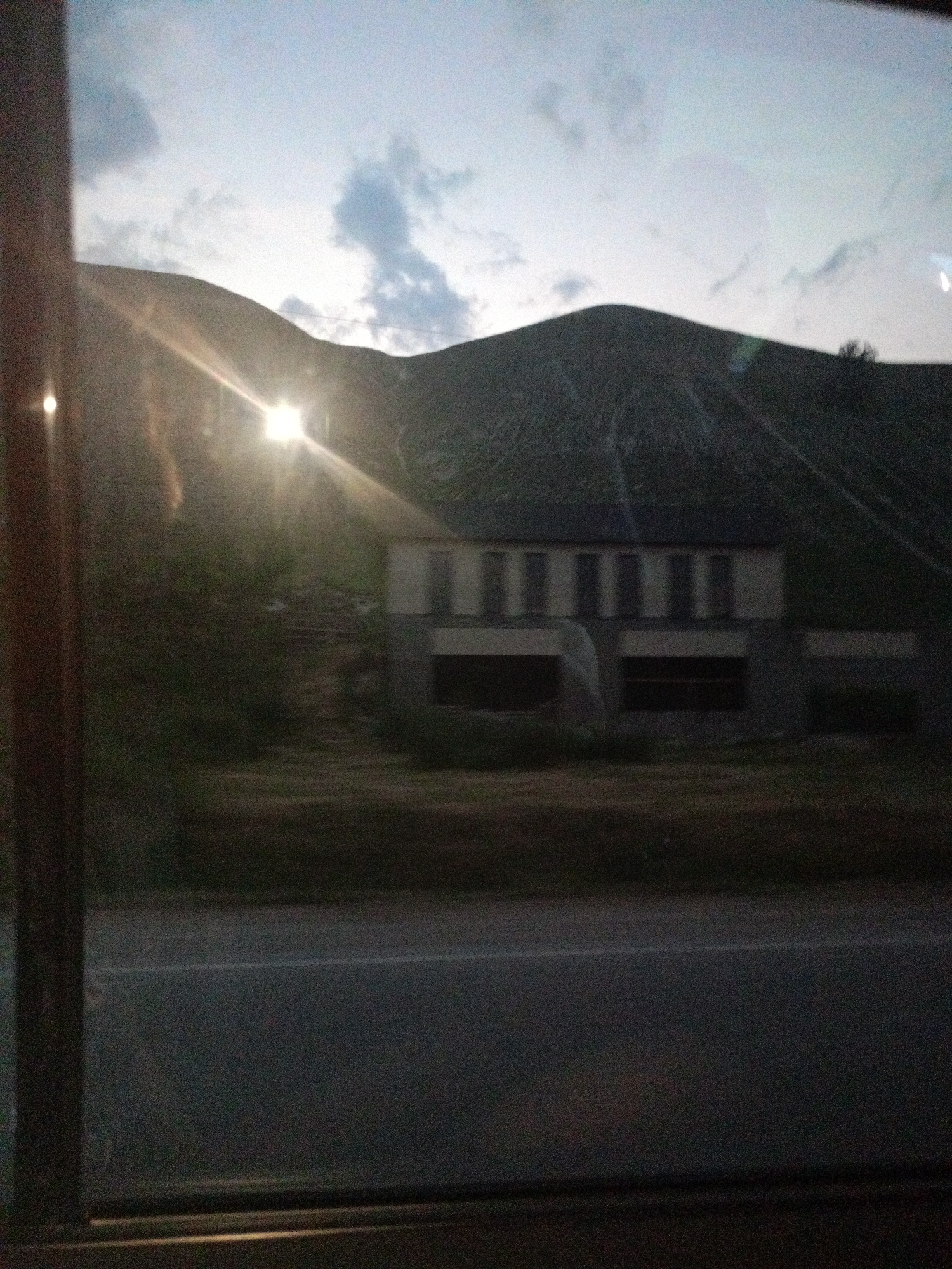 The image depicts a house situated on a hillside under a cloudy sky. The sun is visible through the clouds, creating a bright flare in the upper left portion of the image. The house appears to be a two-story structure with a dark roof and light-colored walls. Several windows are visible, some of which are dark, suggesting they may be covered or unlit. The hillside is covered in vegetation, and there are visible steps leading up to the house. In the foreground, a road is visible, with what appears to be a reflective stripe. The overall scene is dimly lit, possibly indicating dawn or dusk. The location is in the city of Кавказский, Russia.