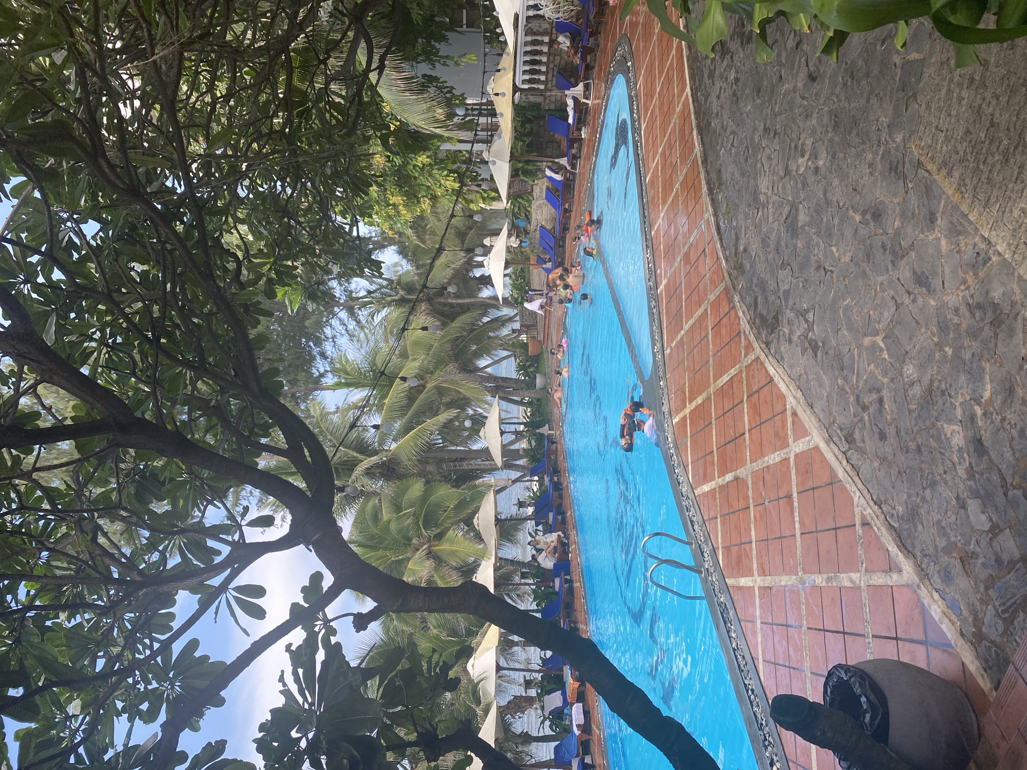 The image depicts a resort swimming pool area in Mũi Né, Vietnam, as evidenced by the tropical foliage including palm trees and frangipani. The foreground features a dark, textured stone pathway leading to a vibrant blue swimming pool with clear water. The pool has a shallow end with metal railings and appears to be populated by several individuals swimming and enjoying the water. The pool area is surrounded by a red tiled deck with decorative stone borders.
Beyond the pool, a row of white sun umbrellas and blue lounge chairs are set up, with some people relaxing on them. In the background, lush green palm trees and other tropical vegetation are visible, with the ocean and a clear blue sky with some clouds completing the scene. The atmosphere suggests a leisurely resort vacation.