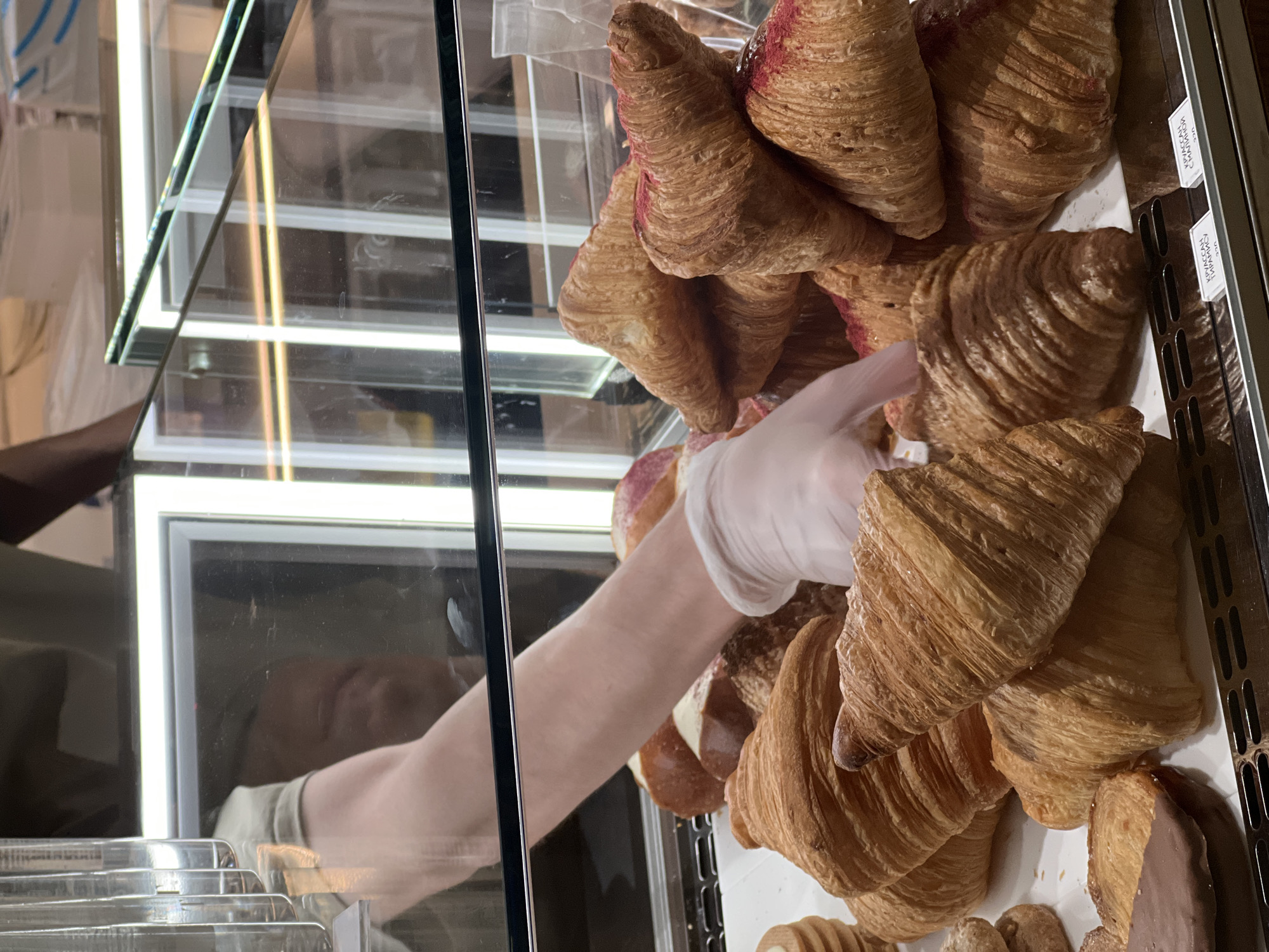 A bakery display case filled with croissants is shown. A person wearing a disposable glove is reaching into the case, possibly to select a pastry. The pastries are displayed in a chaotic manner. The location is in Vladivostok, Russia, as indicated by the Cyrillic text on the pricing labels. The reflection of a person's face can be seen in the glass of the display case. The interior is well-lit.