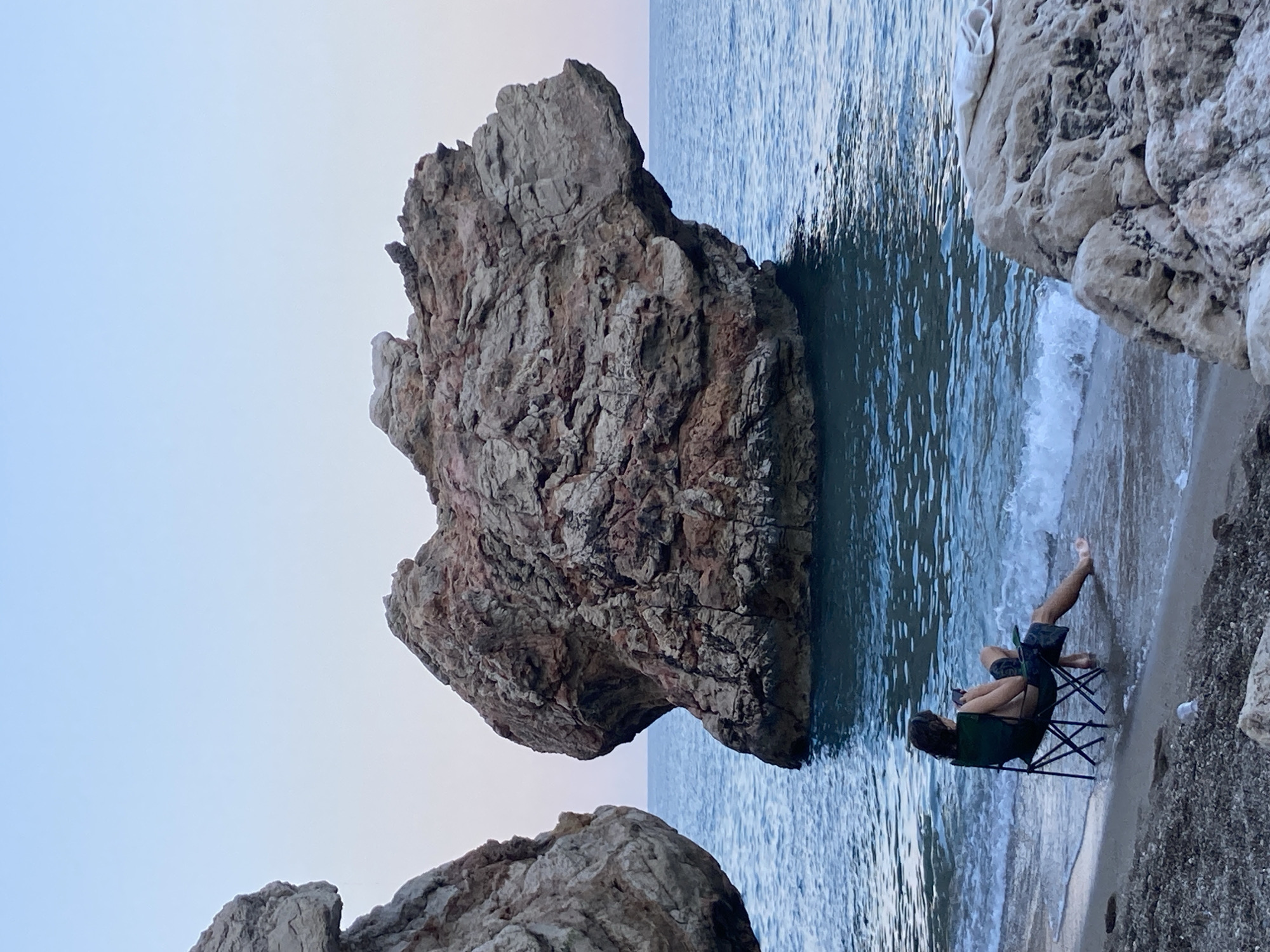 The image depicts a tranquil scene on the coast of Kemer, Turkey. In the foreground, a person is seated in a folding chair at the edge of the water, with their legs submerged. The chair is positioned on a pebble beach. The person is holding a mobile phone. In the background, a large, rugged rock formation rises from the sea. The water is a deep blue, and small waves are lapping at the shore. Two other large rock formations are visible on the left and right sides of the frame, framing the central rock and the sea. The sky above is a clear, pale blue, suggesting either early morning or late evening light. The overall atmosphere is one of peaceful solitude and natural beauty.