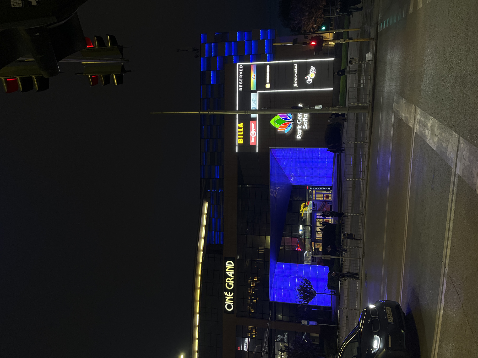 The image captures a vibrant urban street scene at night in Sofia, Bulgaria, centered on a modern shopping complex. The main building, identified by a large sign as "Park Center Sofia," features a contemporary architectural design with prominent blue-lit square panels. Numerous commercial signs are brightly lit, including "CINE GRAND" vertically on the left side, and "BILLA TheOne," "RESERVED," "next LEVEL," "dm," "BURGER KING," and "Scandal" displayed vertically and horizontally on the upper levels.
On the street level, traffic lights show red for vehicles and green for pedestrians, indicating an active intersection. Several people are visible on the sidewalk and near the building's entrance, engaged in typical evening activities like walking or entering the shopping center. A black car with its headlights on is seen on the right, and its Bulgarian license plate "EA 8476 AA" is partially visible. Metal barriers line the pedestrian area. The overall atmosphere is bustling and commercial, characterized by artificial lighting against a dark night sky.