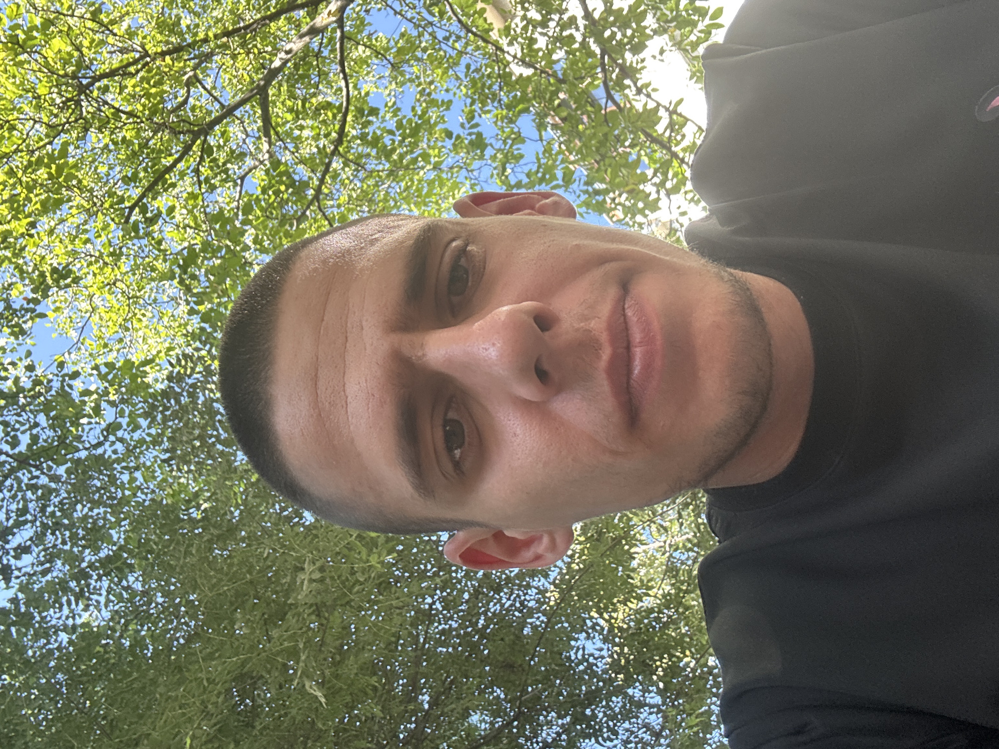 A close-up, low-angle selfie of a man with a buzz cut is taken outdoors, with a bright blue sky and green foliage from trees visible in the background. The man's face is centered in the frame, with his eyes open and looking directly at the camera. He is wearing a black t-shirt. The lighting suggests it is daytime. The specific location in Sofia, Bulgaria is not determinable from the provided image.