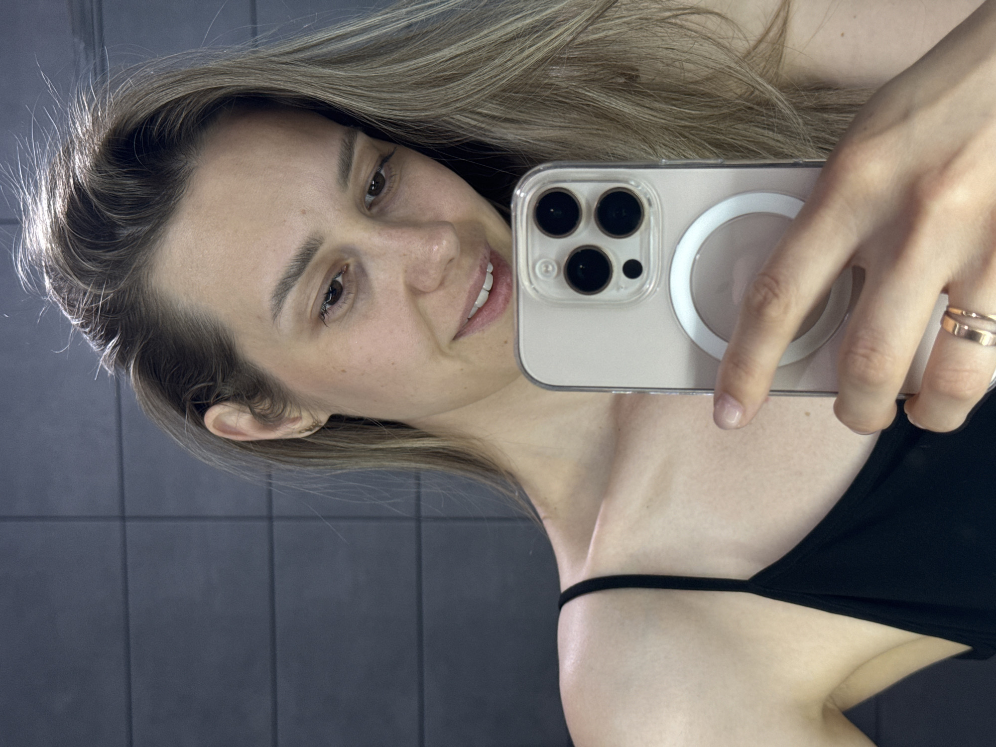 A woman is taking a selfie with a smartphone. The phone has a transparent case with a magnetic ring. She is wearing a black tank top. The background appears to be a tiled wall, possibly in a bathroom. There's no indication of the location as the city of Chaikovsky, Russia.