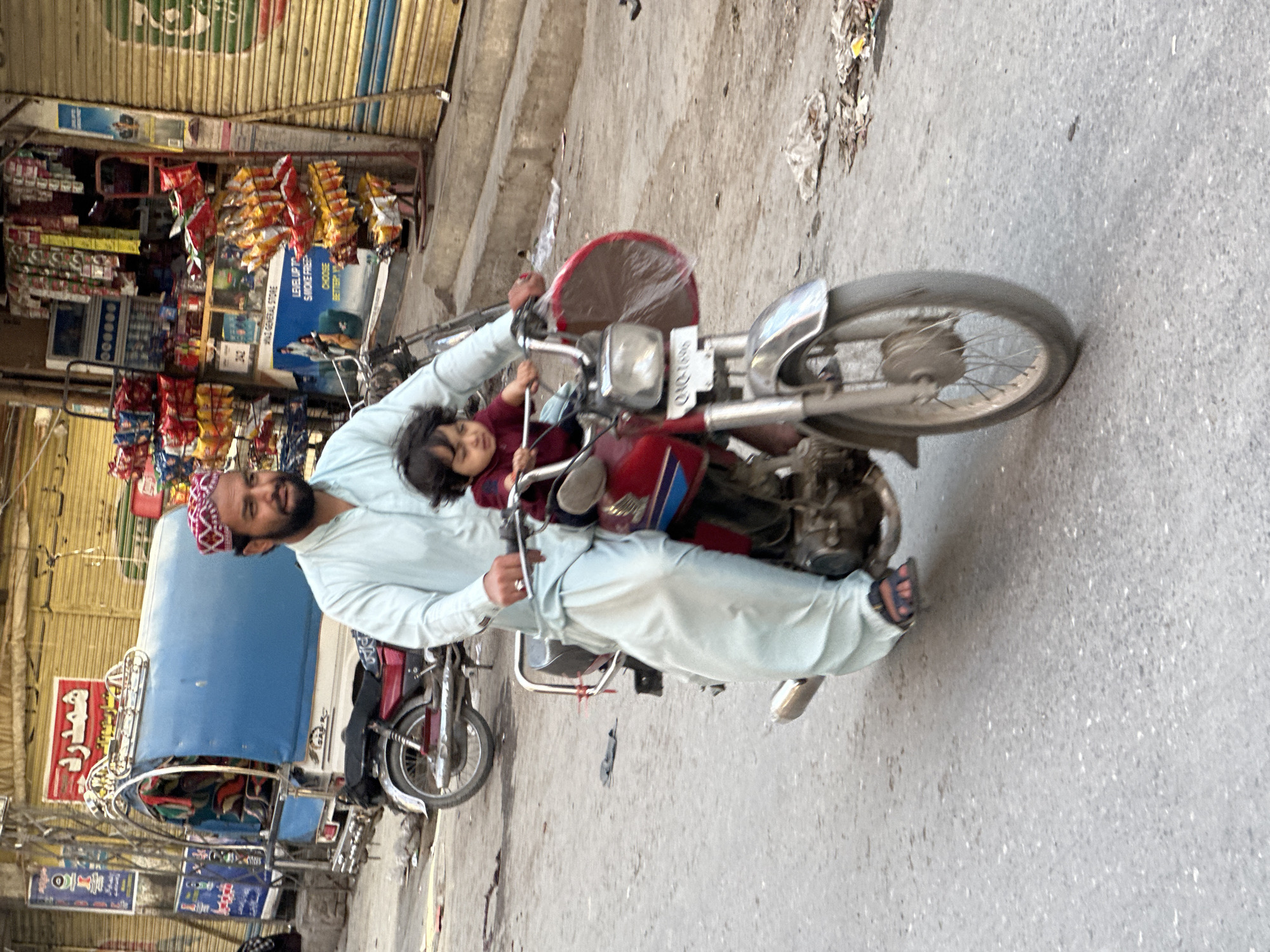 Here is a description of the image:
The image captures a scene on a street in what appears to be Quetta City Tehsil, Pakistan. A man with a beard and wearing a traditional red and white cap is seated on a red motorcycle, with a small child in front of him, between his arms. The motorcycle has a license plate that reads "QAQ-1696". To the left of the motorcycle, there is a blue auto-rickshaw. The background reveals a commercial area with shops displaying a variety of products, including snacks and packaged goods. One shop appears to be named "WAZ GENERAL STORE". The street is paved and shows signs of litter. The buildings in the background have corrugated metal exteriors.