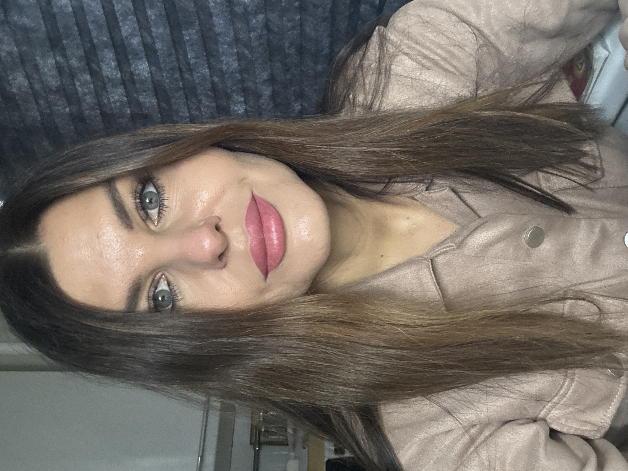 A woman with light skin, light green eyes, and medium-brown hair poses for a selfie. She wears a light brown jacket. The background is a dark gray, vertically-lined surface. The setting is unknown, and there is no verifiable evidence to place the subject in Chaikovsky, Russia.