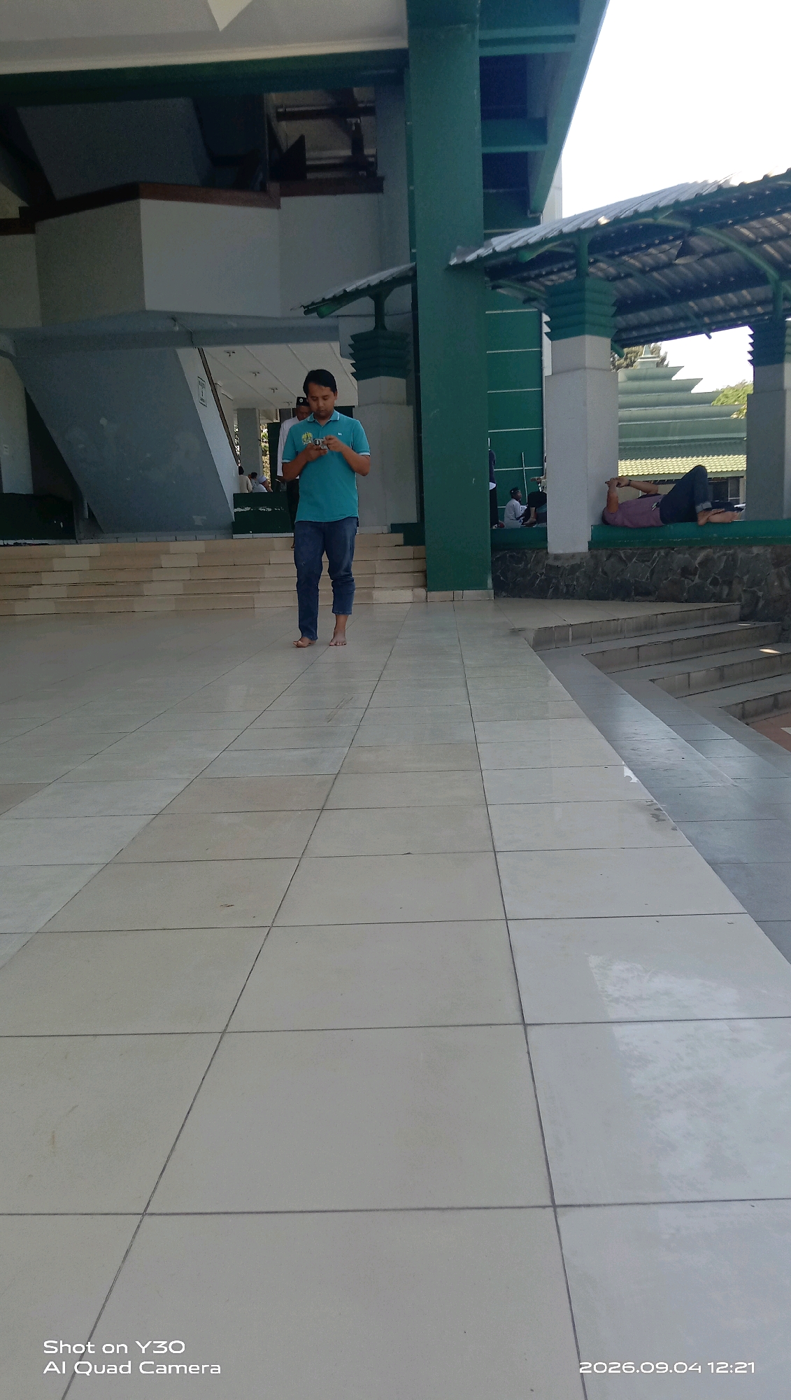 In Klangonan, Indonesia, a young man in a teal polo shirt and blue jeans stands barefoot on a tiled walkway. He is looking down at a mobile phone in his hands. Behind him, a tiered, green-and-white building with a covered walkway is visible. To the right, another individual is lying down on a raised platform. The walkway is made of light-colored square tiles.