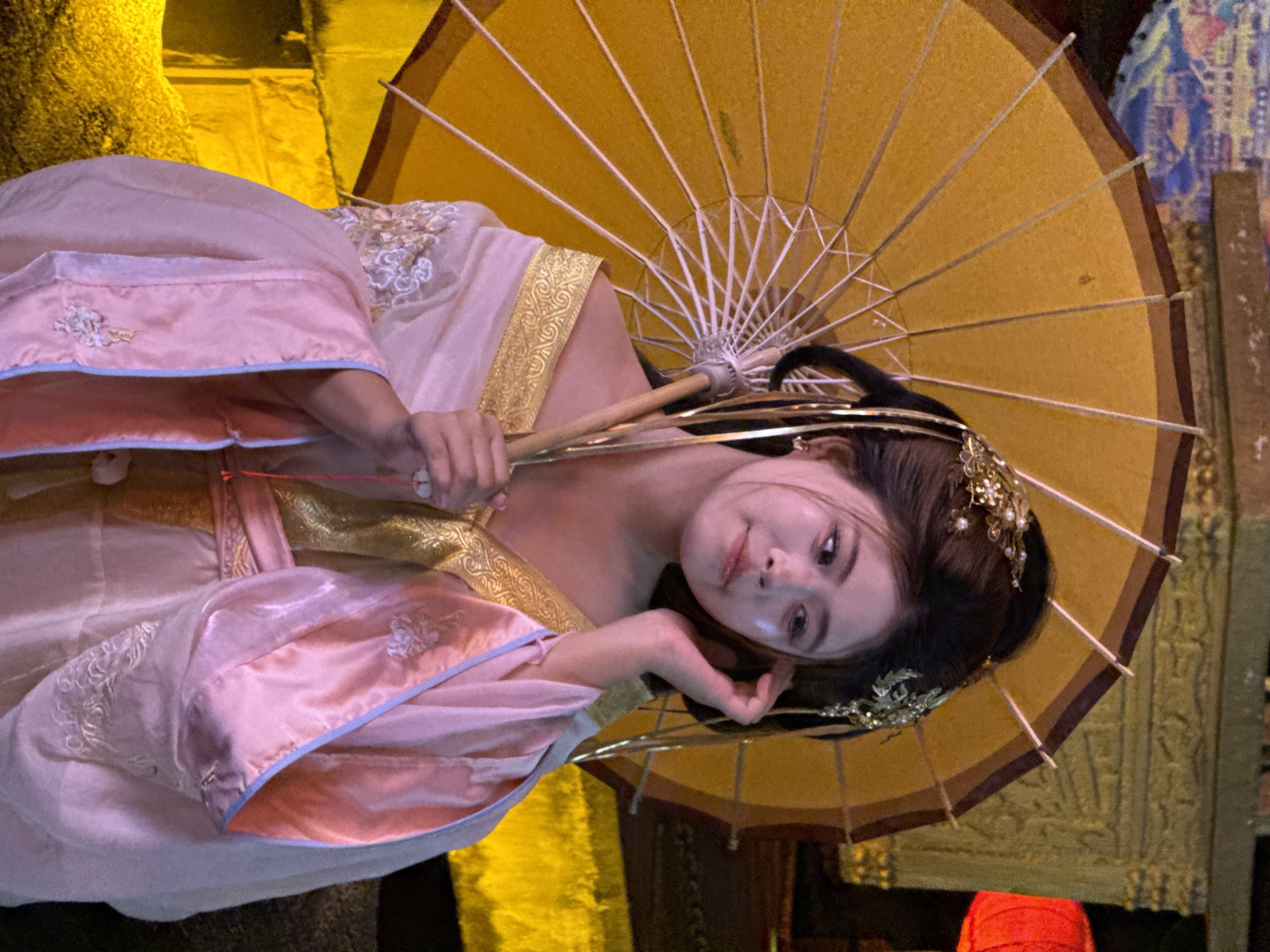 A woman is posing for a photo under a yellow umbrella. She is wearing a pink and gold garment, and her hair is adorned with a jeweled headpiece. The setting appears to be a street in Yuzhong District, China.