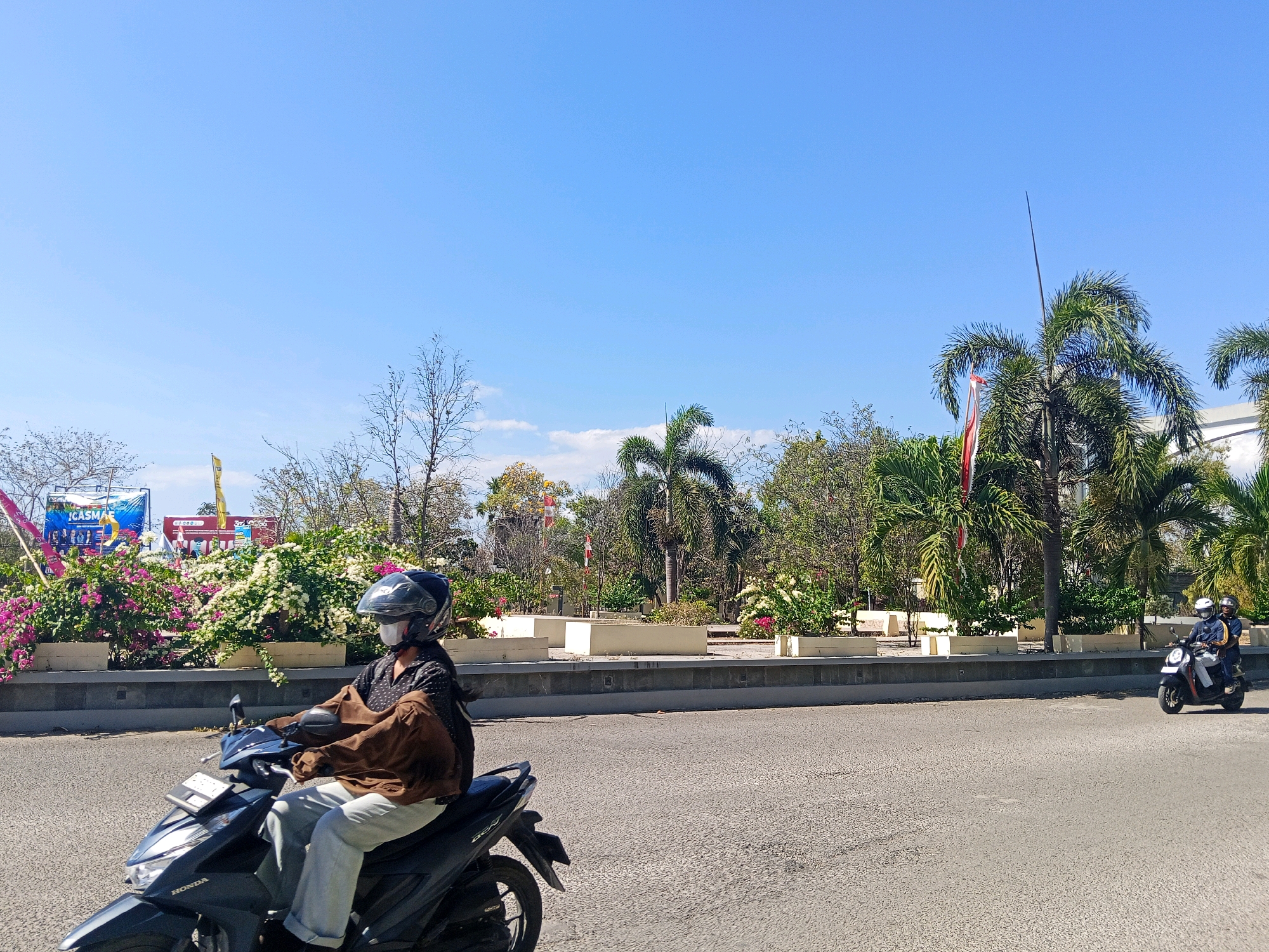 The image depicts a street scene in Kupang, Indonesia, under a clear blue sky. In the foreground, a person wearing a helmet and a mask rides a black Honda scooter. They are dressed in a brown jacket and light-colored pants. The scooter has a license plate holder and mirrors. Further down the road, another scooter carrying two people, also wearing helmets and masks, is visible.
The background features a landscaped area with a row of white planter boxes filled with flowering bushes in shades of pink and white. Beyond the planters, various trees, including palm trees, are present. Banners with text are displayed on the left side of the image. The road surface is asphalt. The overall impression is a sunny day in an urban environment.