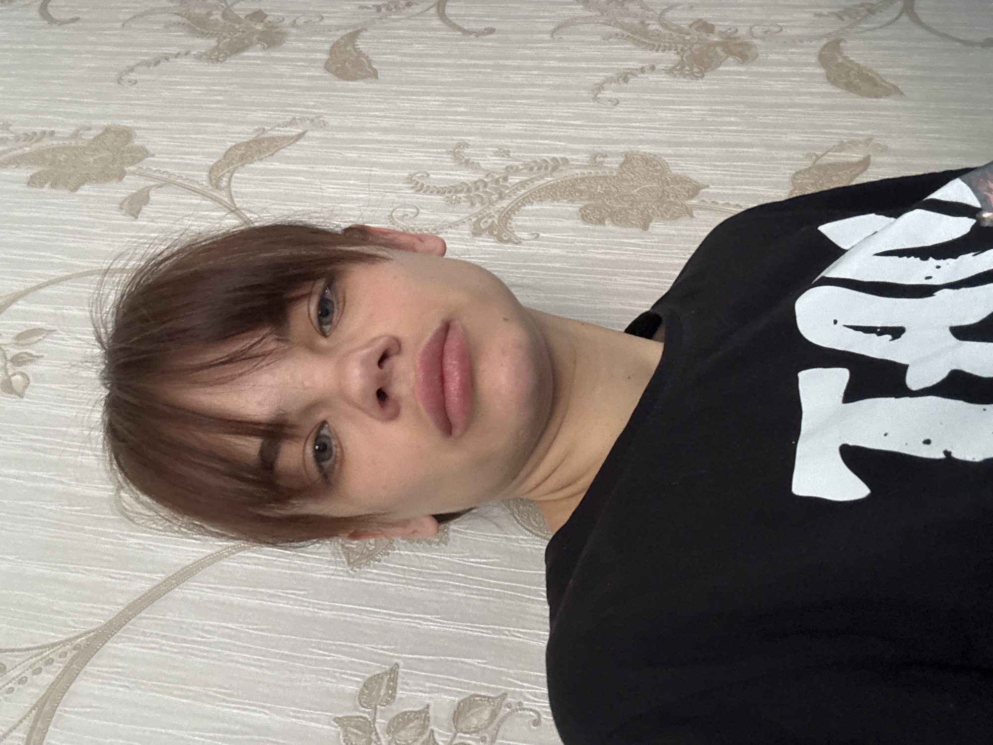 The image features a close-up of a young person lying down, with their head tilted to the left. They have light brown hair styled with bangs covering their forehead, fair skin, and blue-grey eyes, which are looking directly at the camera. Their expression is neutral, with slightly parted lips.
The individual is wearing a plain black t-shirt. On the front of the t-shirt, there is a large, prominent white graphic that appears to be stylized text or characters. While specific words are not clearly legible, the shapes suggest a non-Latin script, potentially Japanese or a similar artistic font.
The background is a light-colored surface, likely wallpaper or bedding, adorned with a repeating pattern of intricate, muted beige floral and ornamental designs. This patterned setting indicates an indoor environment, such as a bedroom or living area. The lighting is even, typical of indoor illumination. The overall scene is a candid portrait, possibly a selfie, within a residential space in Rostov-on-Don, Russia.