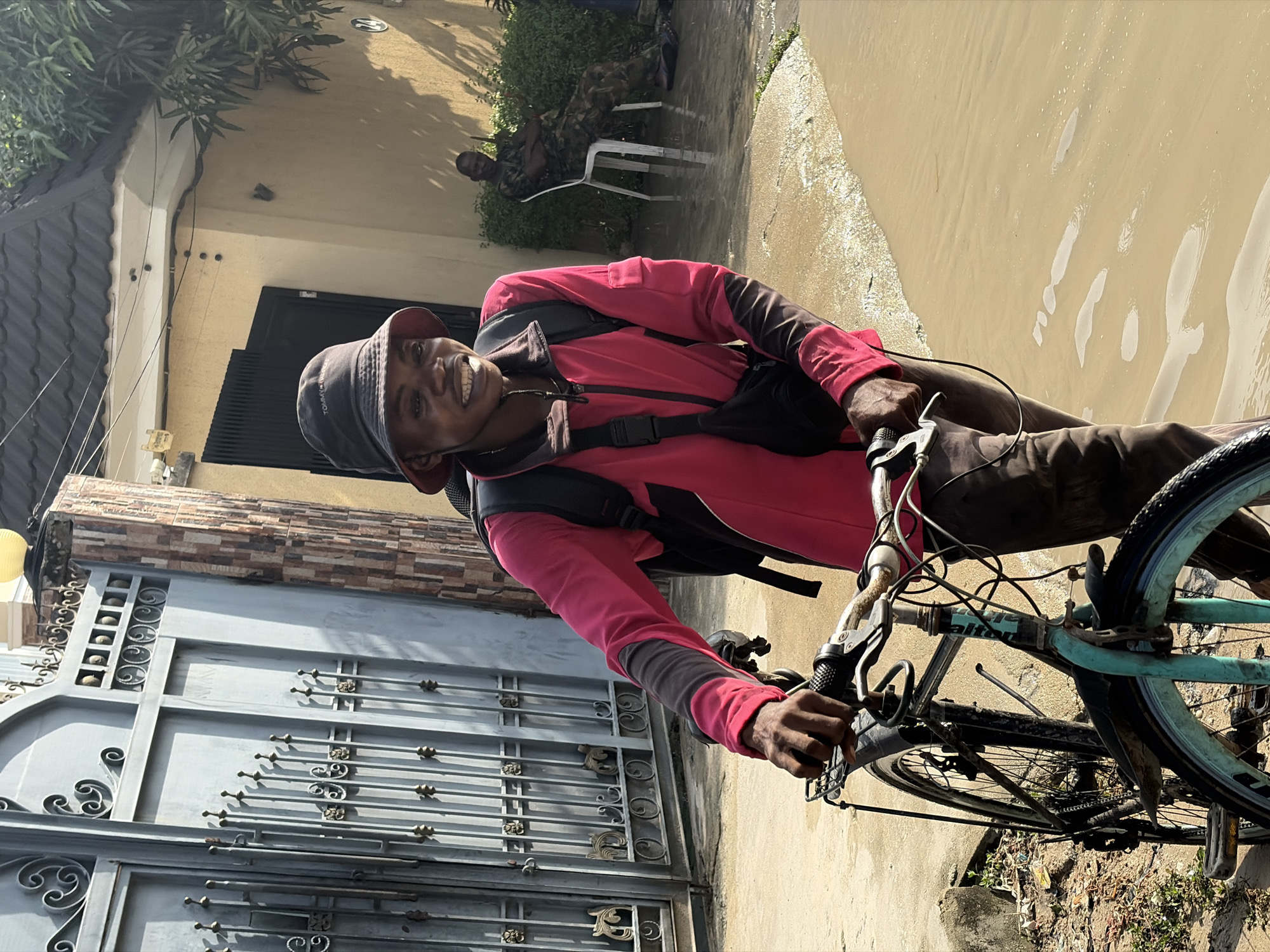 A man is riding a bicycle in Igboefon, Nigeria. He is wearing a red jacket, brown pants, and a bucket hat. He has a black backpack on his back. The bicycle is a light blue color. Behind him is a metal gate and a building with a tan wall and a dark gray roof. In the background, a man in military fatigues is sitting in a white plastic chair. The ground is paved, and there is some foliage visible. The lighting suggests it is daytime.