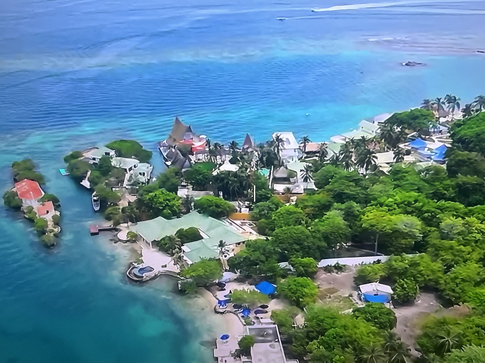 The image is an aerial view of a tropical coastal area with clear blue water. Numerous buildings, varying in size and style, are scattered throughout the landscape, interspersed with lush green trees and vegetation. Several structures have distinctive pointed roofs, some thatched, suggesting a resort or residential development. The coastline features sandy beaches and rocky outcrops. In the water, a few boats are visible, and there appear to be swimming pools or water features associated with some of the buildings. The scene conveys a sense of a secluded and scenic location, likely a tourist destination.