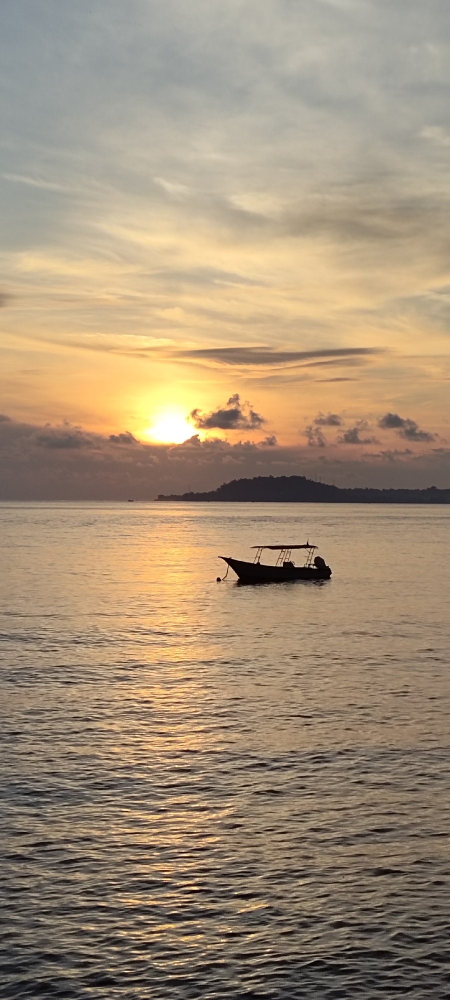 A serene sunset paints the sky in hues of orange and yellow over a calm sea. A solitary boat, silhouetted against the vibrant sky, floats gently on the water. In the distance, the silhouette of a lush, hilly island is visible, with faint structures suggesting human presence. The water reflects the warm glow of the setting sun, creating shimmering patterns on its surface. The overall atmosphere is tranquil and picturesque, capturing a moment of natural beauty in Sabang, Indonesia.